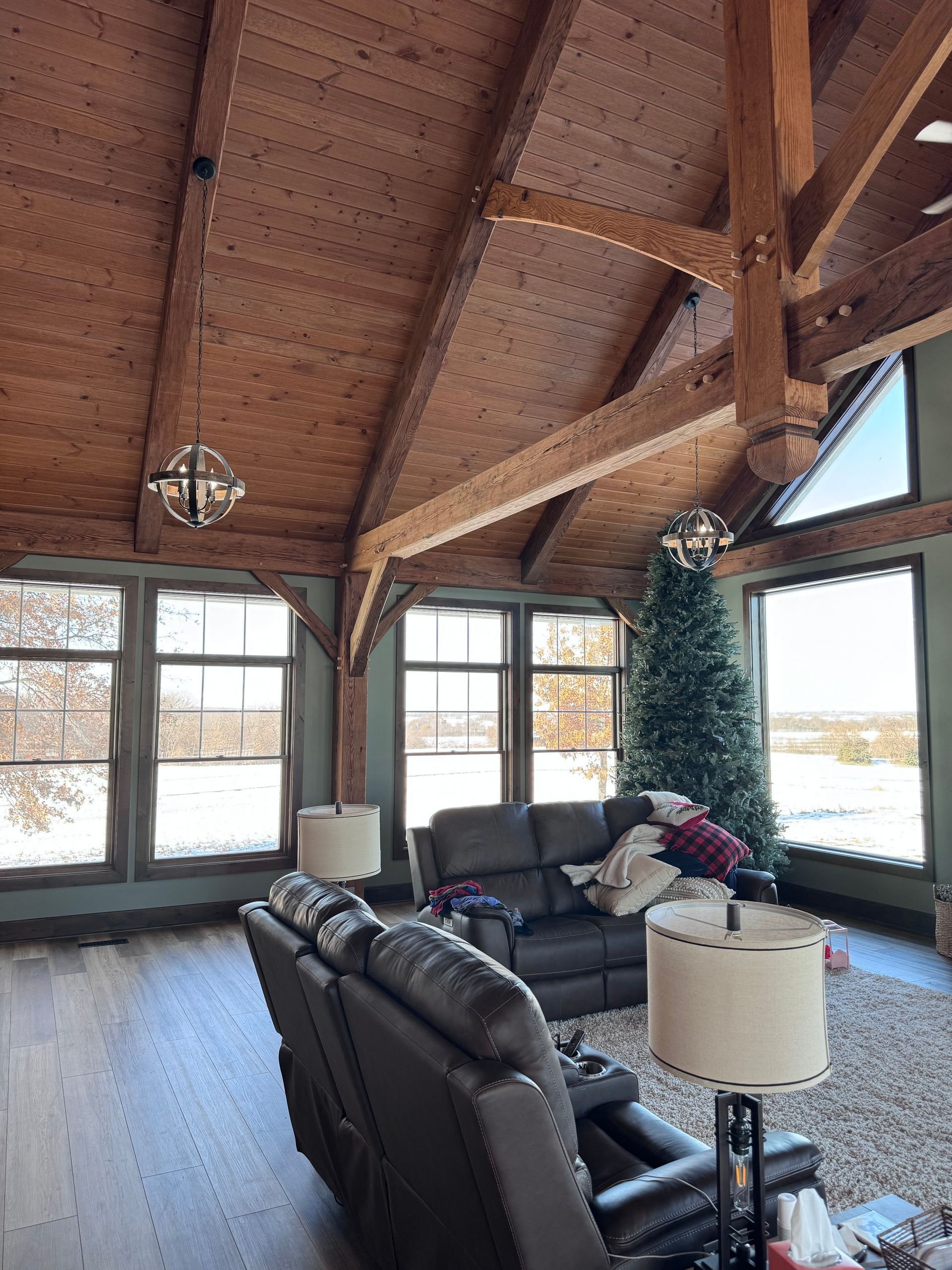 Living room with high wooden ceiling, large windows, dark grey sofa, Christmas tree, and spherical chandeliers.