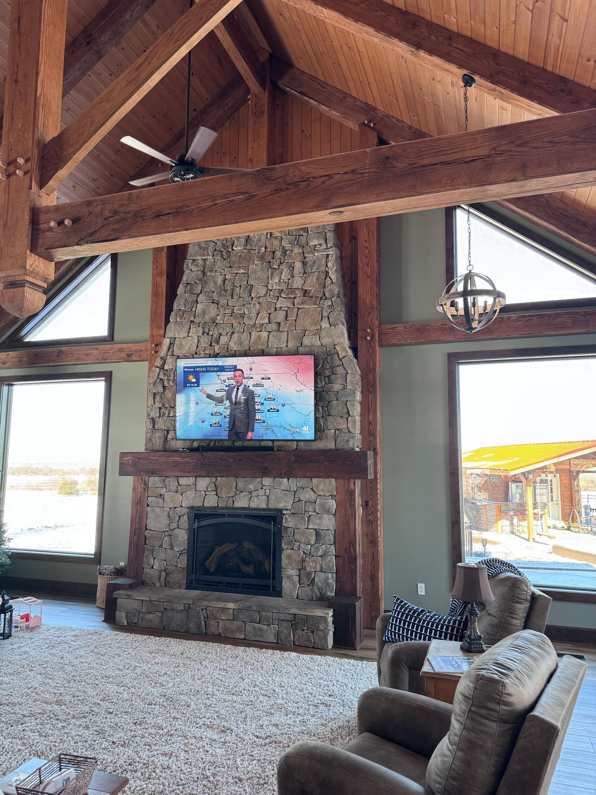 Stone fireplace with TV and wooden beams in a cabin-style living room. Large windows, two armchairs, and a ceiling fan.