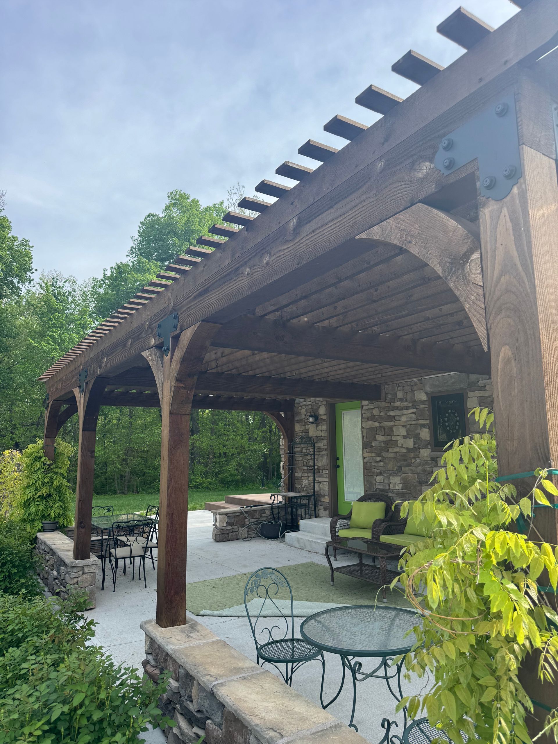 Wooden pergola over a patio with a stone wall and seating. Trees in the background.