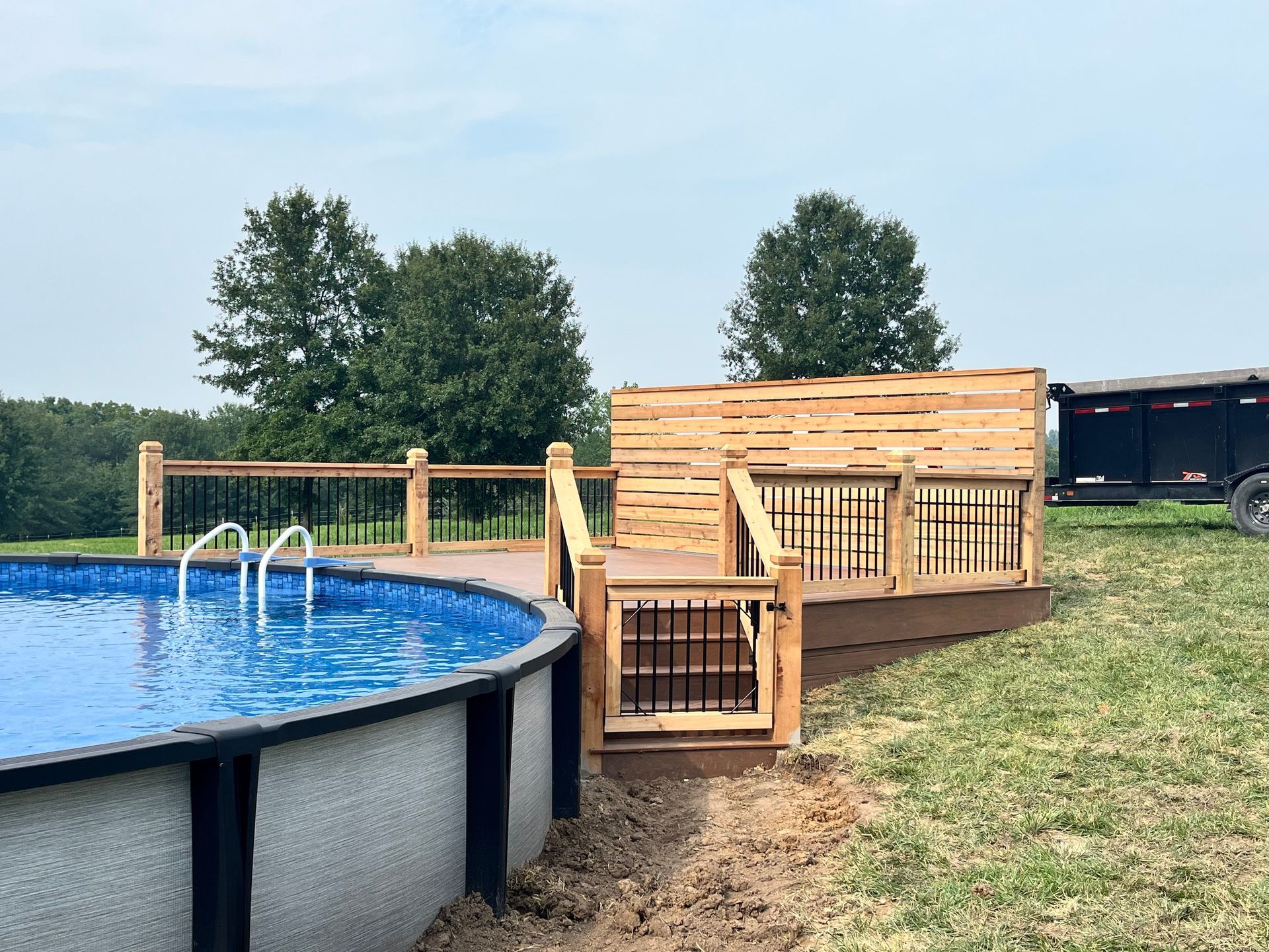 Above-ground pool with wooden deck and fence, dark railing, and steps. Trees and grass in the background.
