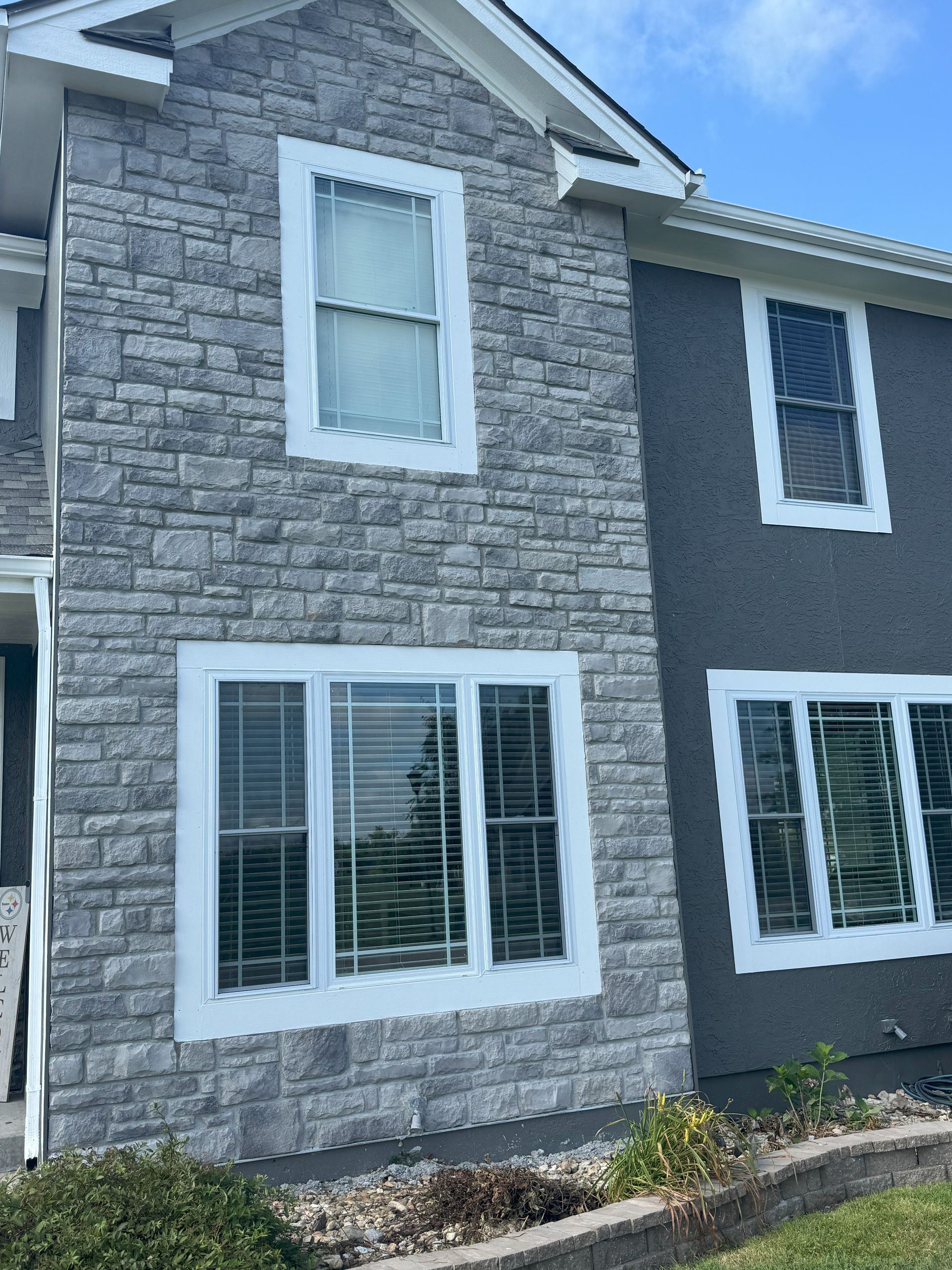 Two-story house exterior with stone and stucco siding, white window frames, and blue sky.