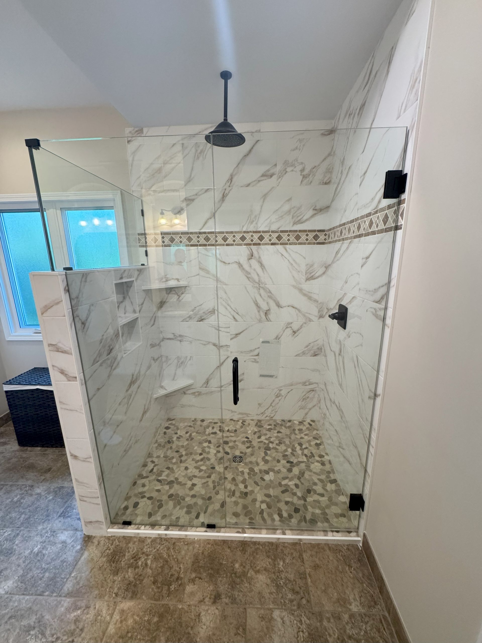 Modern shower with marble-like tile, pebble floor, and glass enclosure. Includes shelves and dark fixtures.