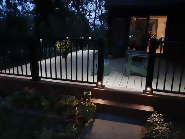 Black deck railing with built-in lights illuminates the wood deck at night, with house visible in the background.