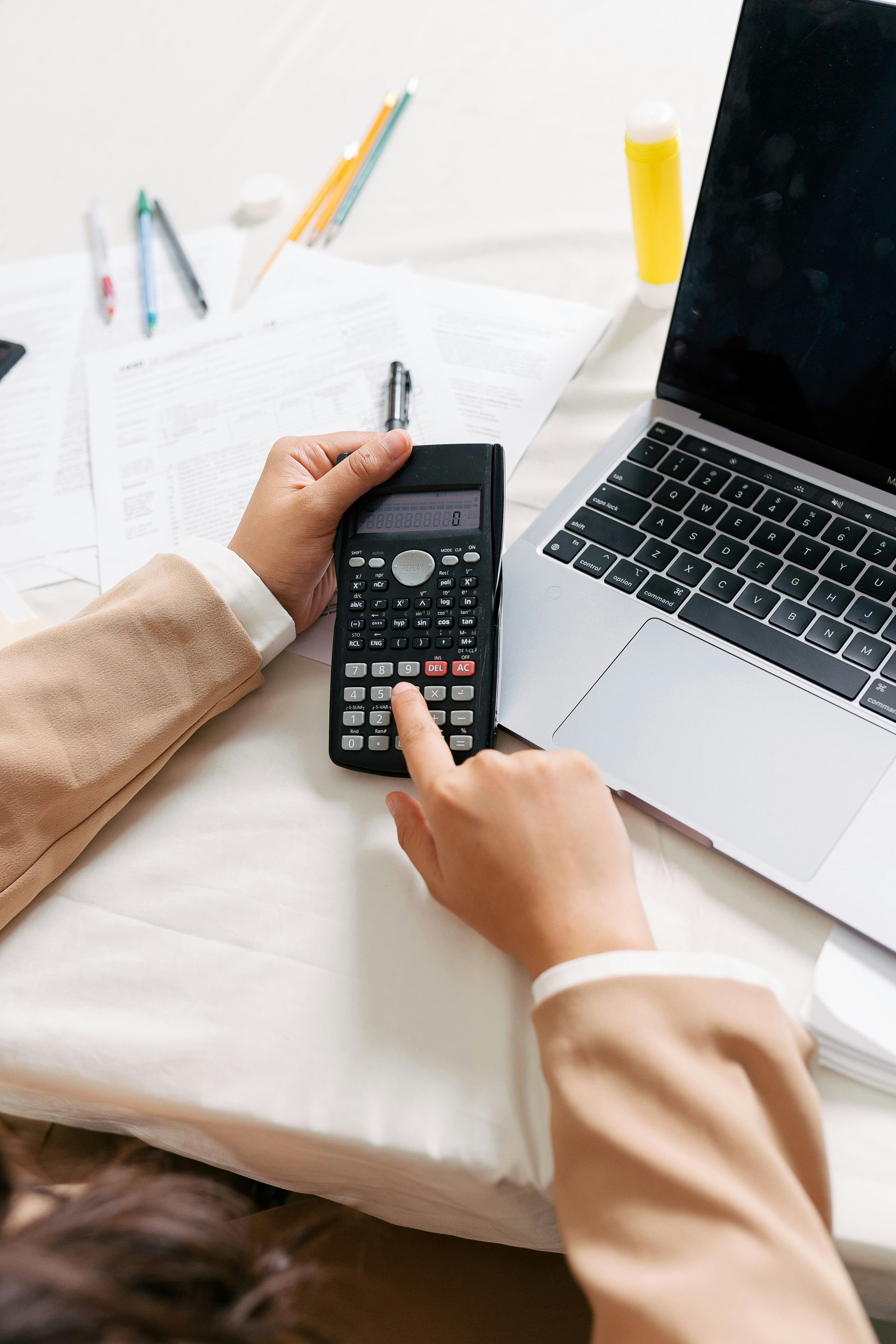 Person using a scientific calculator on a white surface with laptop, papers, and pens.