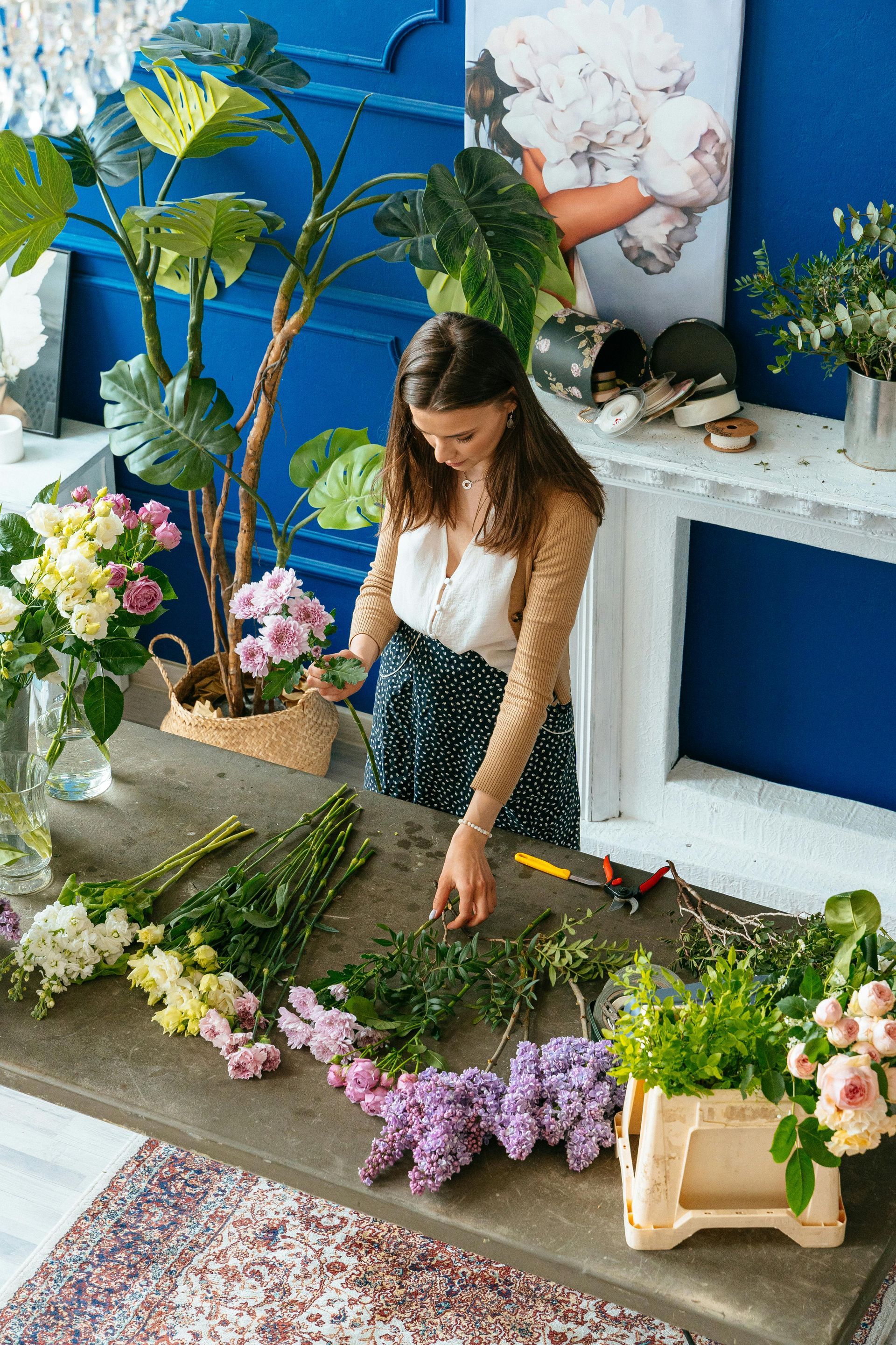 Woman arranging flowers at a wooden table, blue wall background, bright flowers.