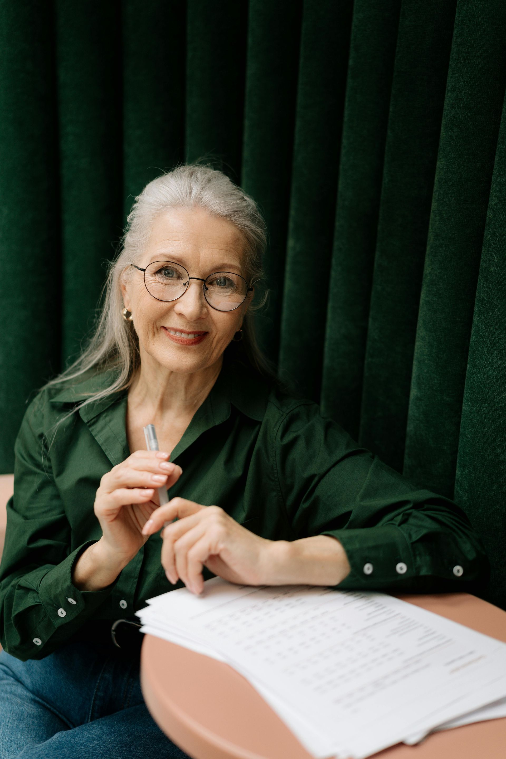 Woman in glasses, holding pen, smiles at camera. Sitting at a table with papers, against a green background.