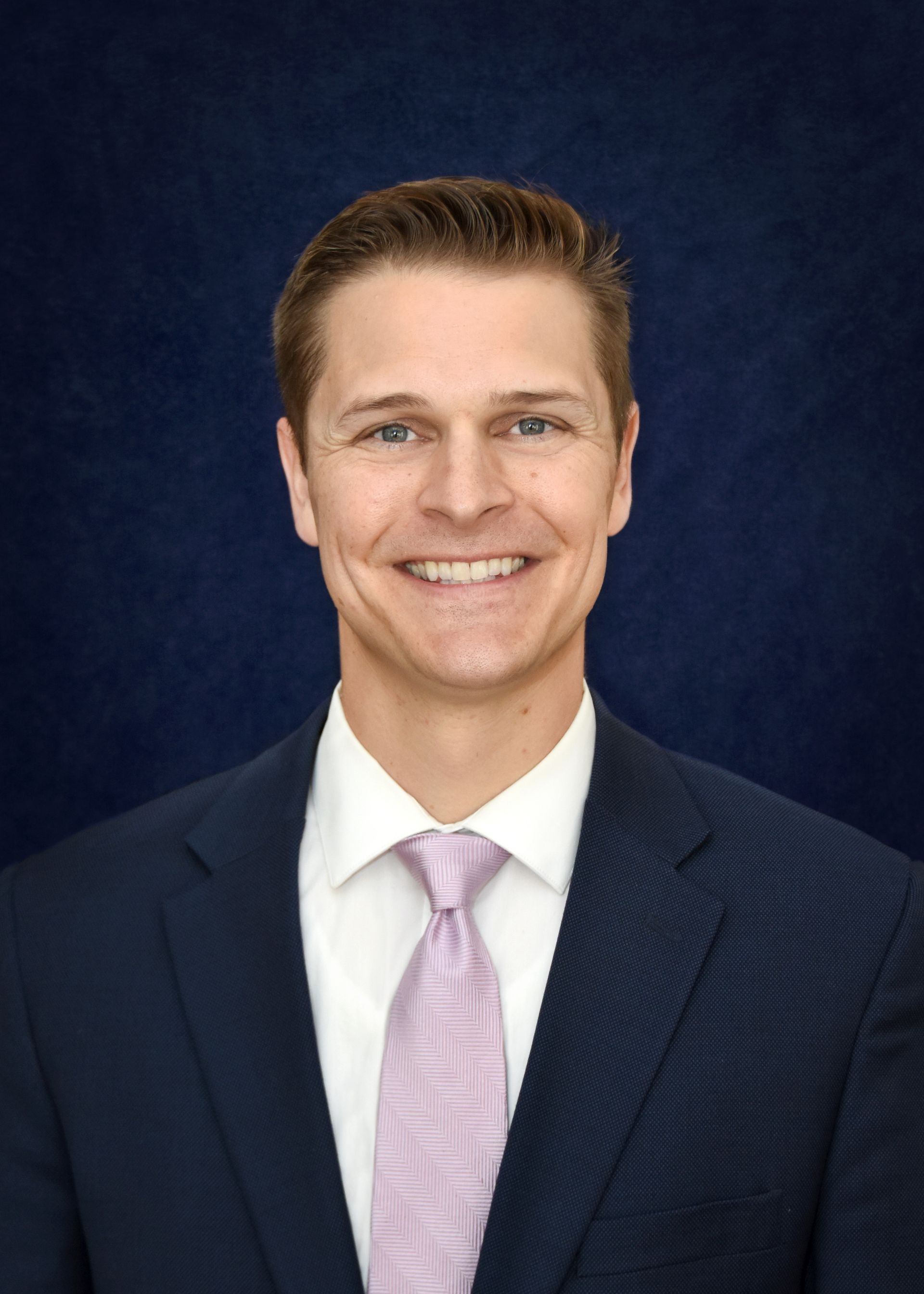 Man in navy suit, white shirt, and pink tie smiles against a blue background.