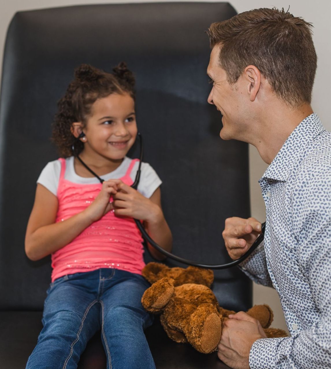 Girl using stethoscope on a teddy bear, smiling at a doctor in an exam room.