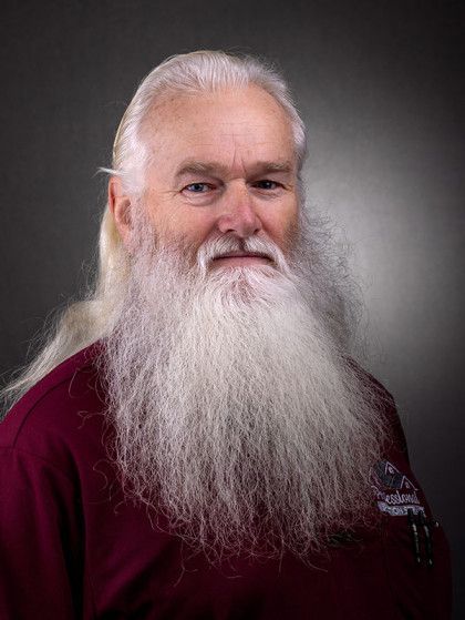 Portrait of a smiling person with long white beard and hair, wearing a maroon shirt against a gray background