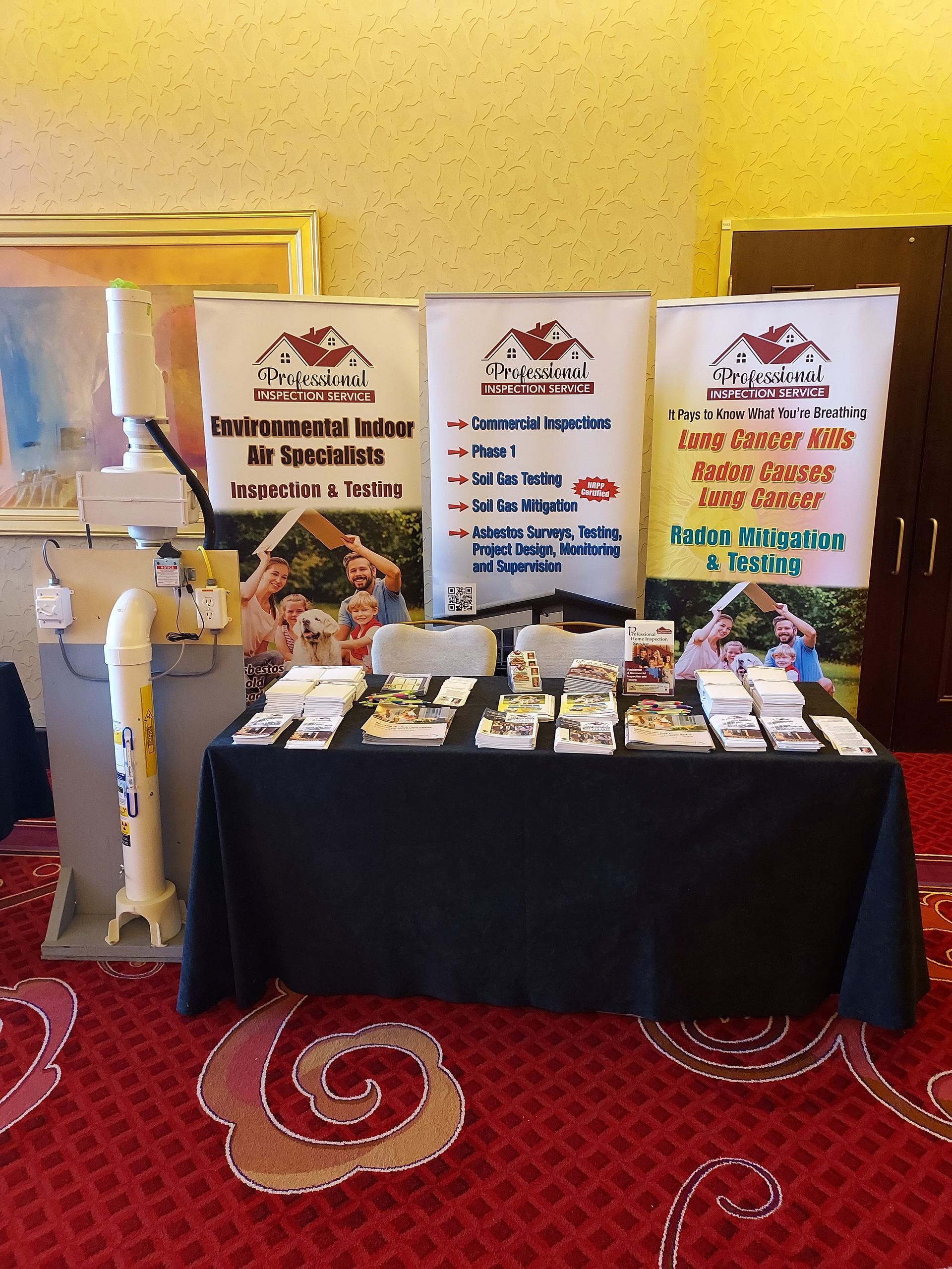 Trade show booth with black table, banners, brochures, and a mannequin beside a cream wall and red carpet.