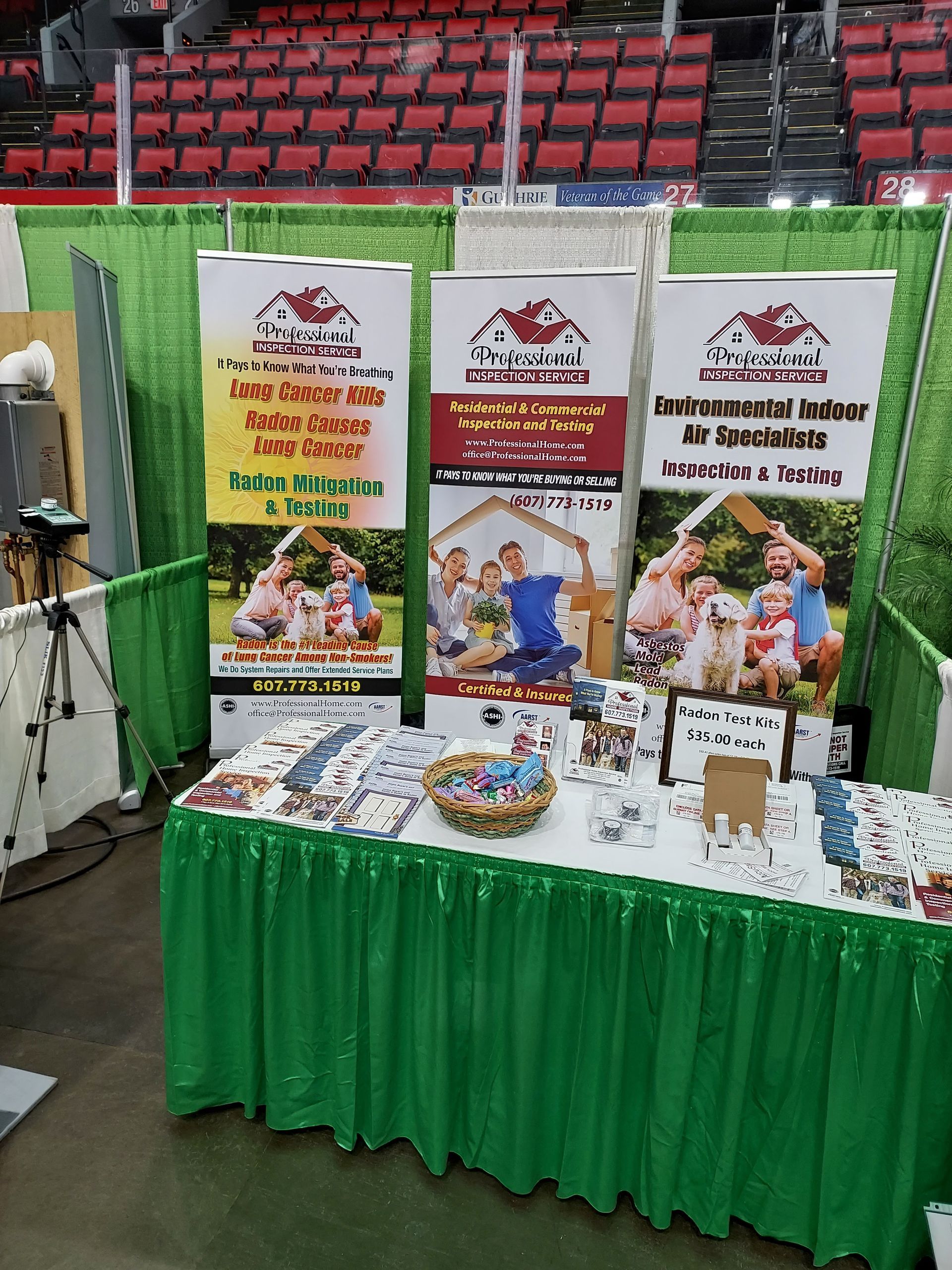Indoor booth with green tablecloths and banners displaying brochures and posters in a stadium setting