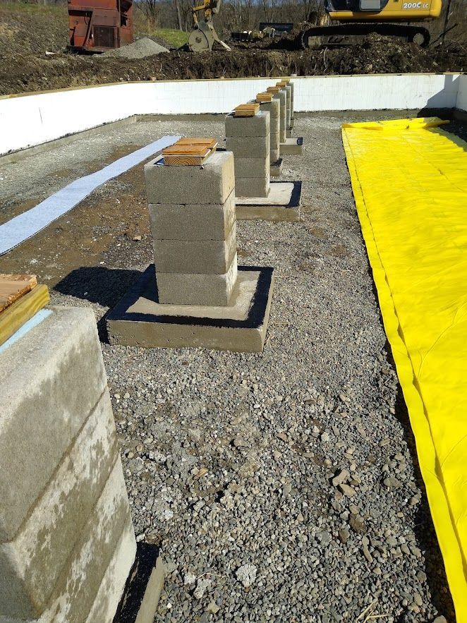 Construction site with stacked concrete blocks on gravel beside white foundation walls and yellow sheeting.
