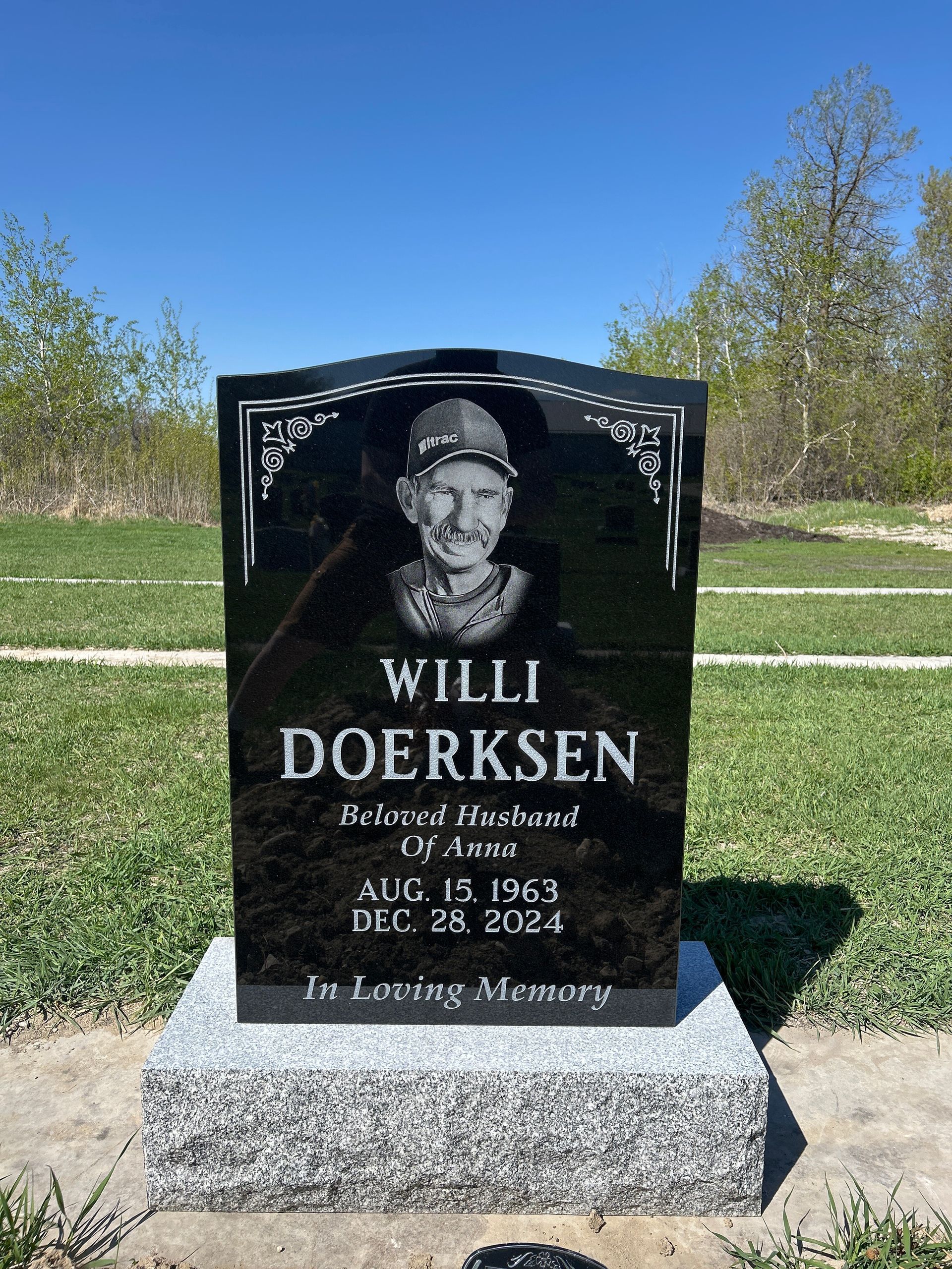 Black memorial gravestone with portrait and engraved text in a sunny cemetery.