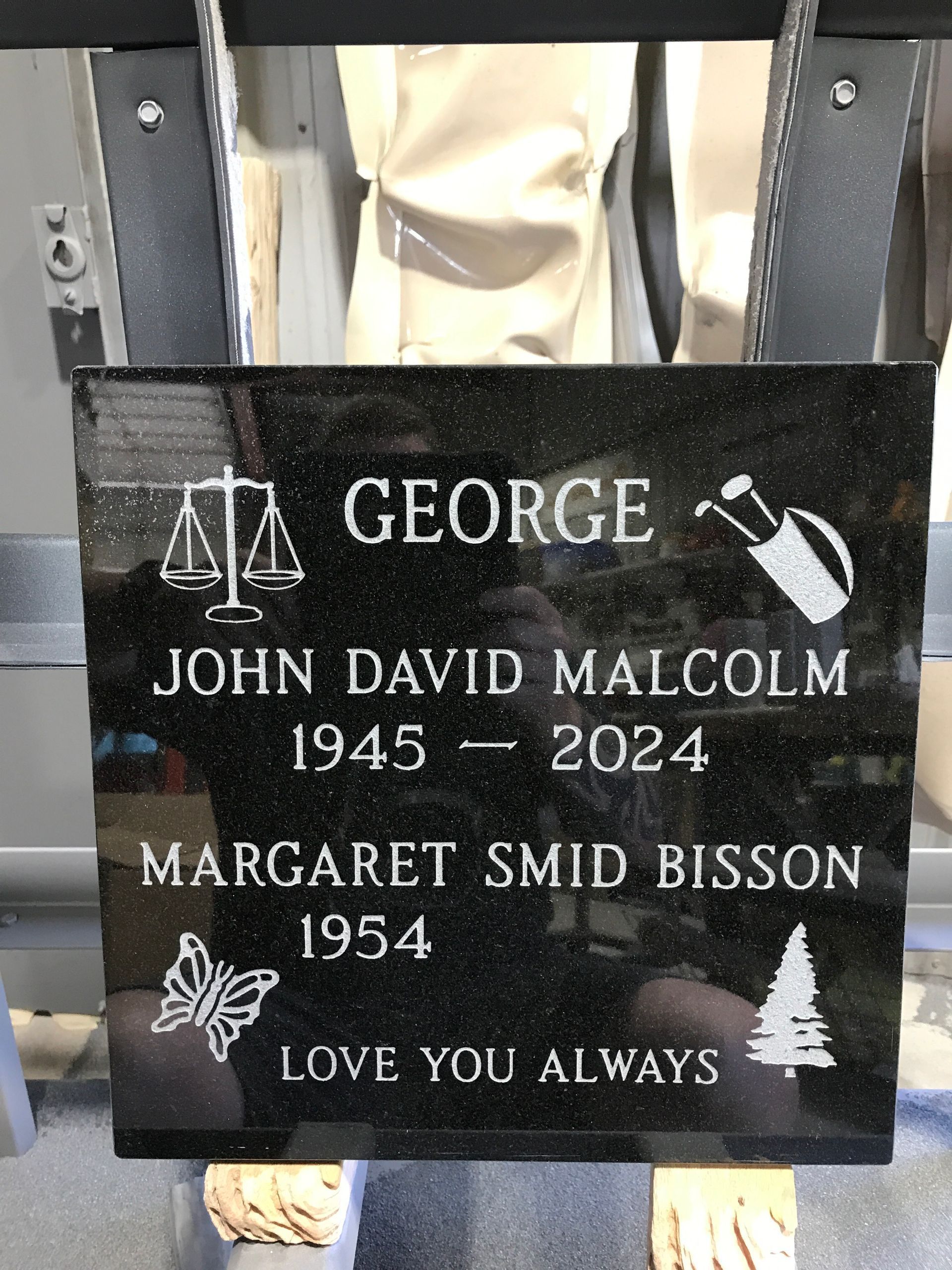 Black memorial plaque with white text: George, John David Malcom 1945–2024, Margaret Smid Bisson 1954–, Love You Always