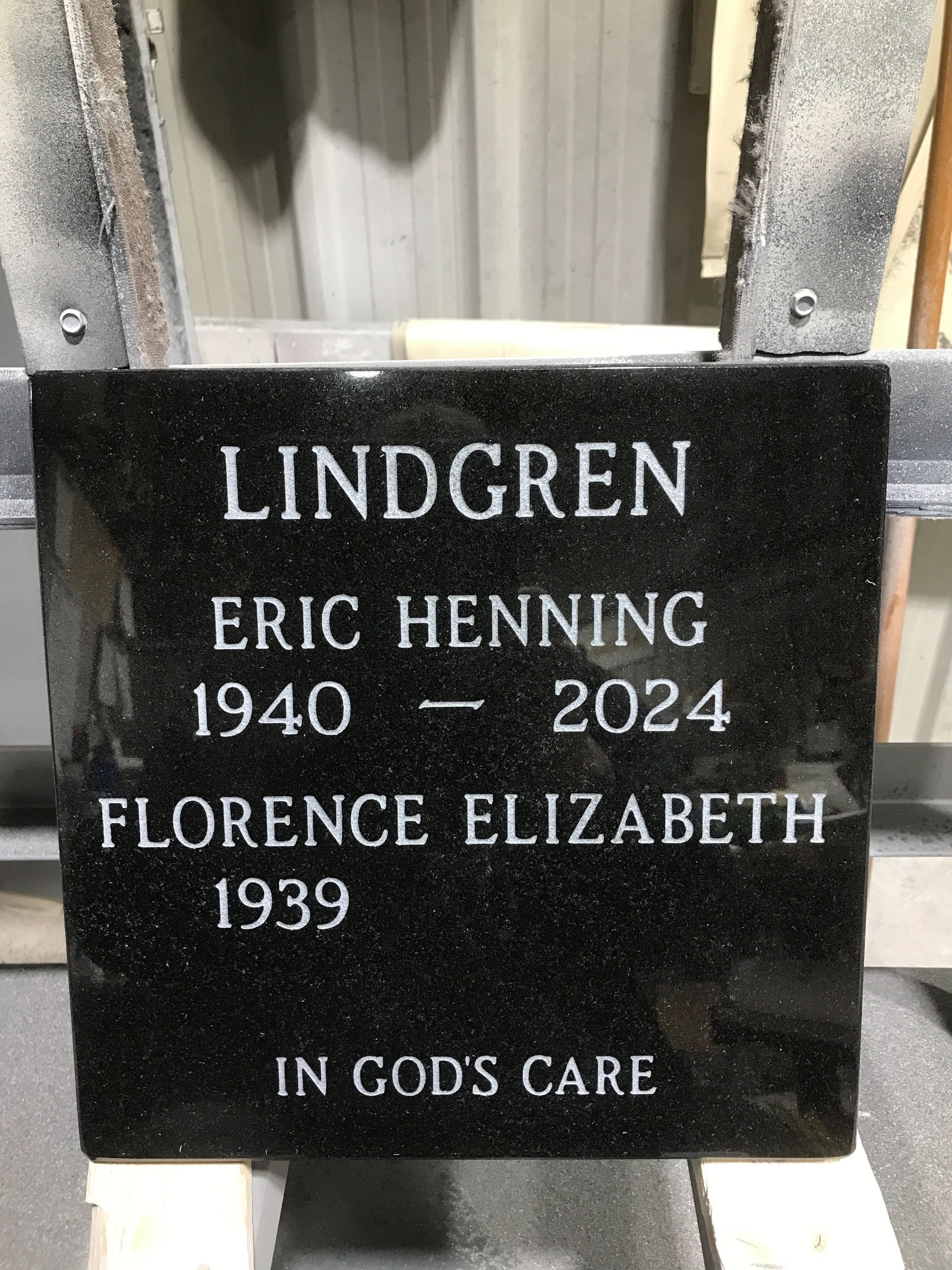 Black memorial headstone engraved “LINDGREN” with names and dates, set outdoors by a fence.