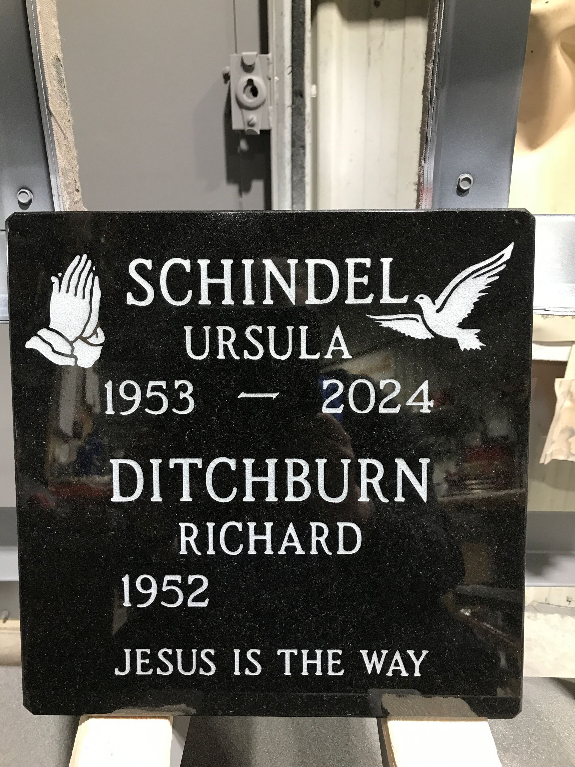 Black memorial plaque with dove, reading “Schindel Ursula 1953–2024” and “Ditchburn Richard 1952,” with “Jesus is the way.”