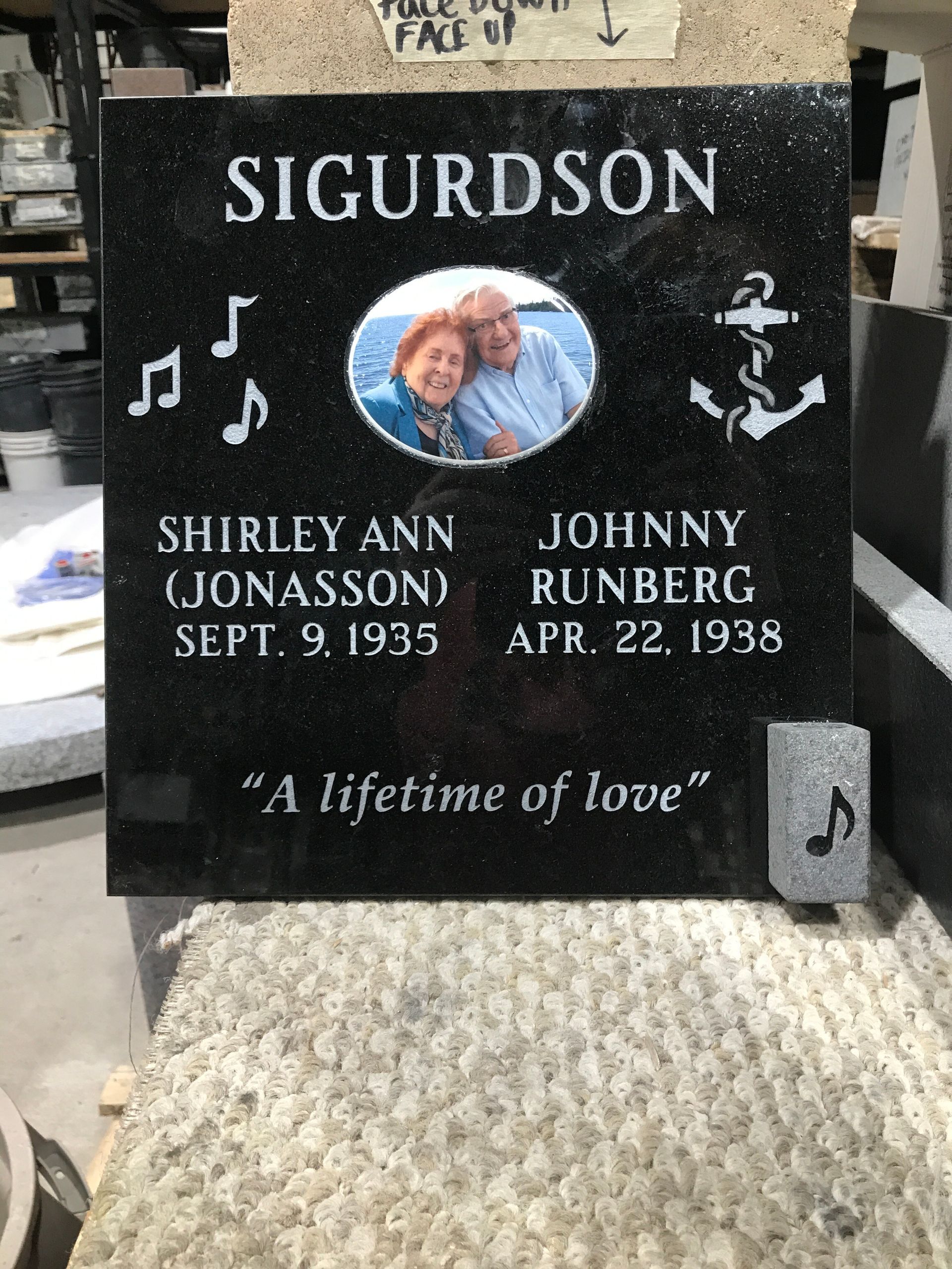 Black granite memorial marker with oval photo, engraved names, dates, musical notes, and anchor symbols.