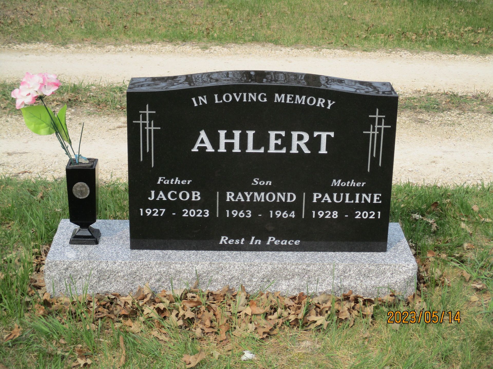 Black memorial headstone with flowers, engraved “Ahlert,” in a grassy cemetery.