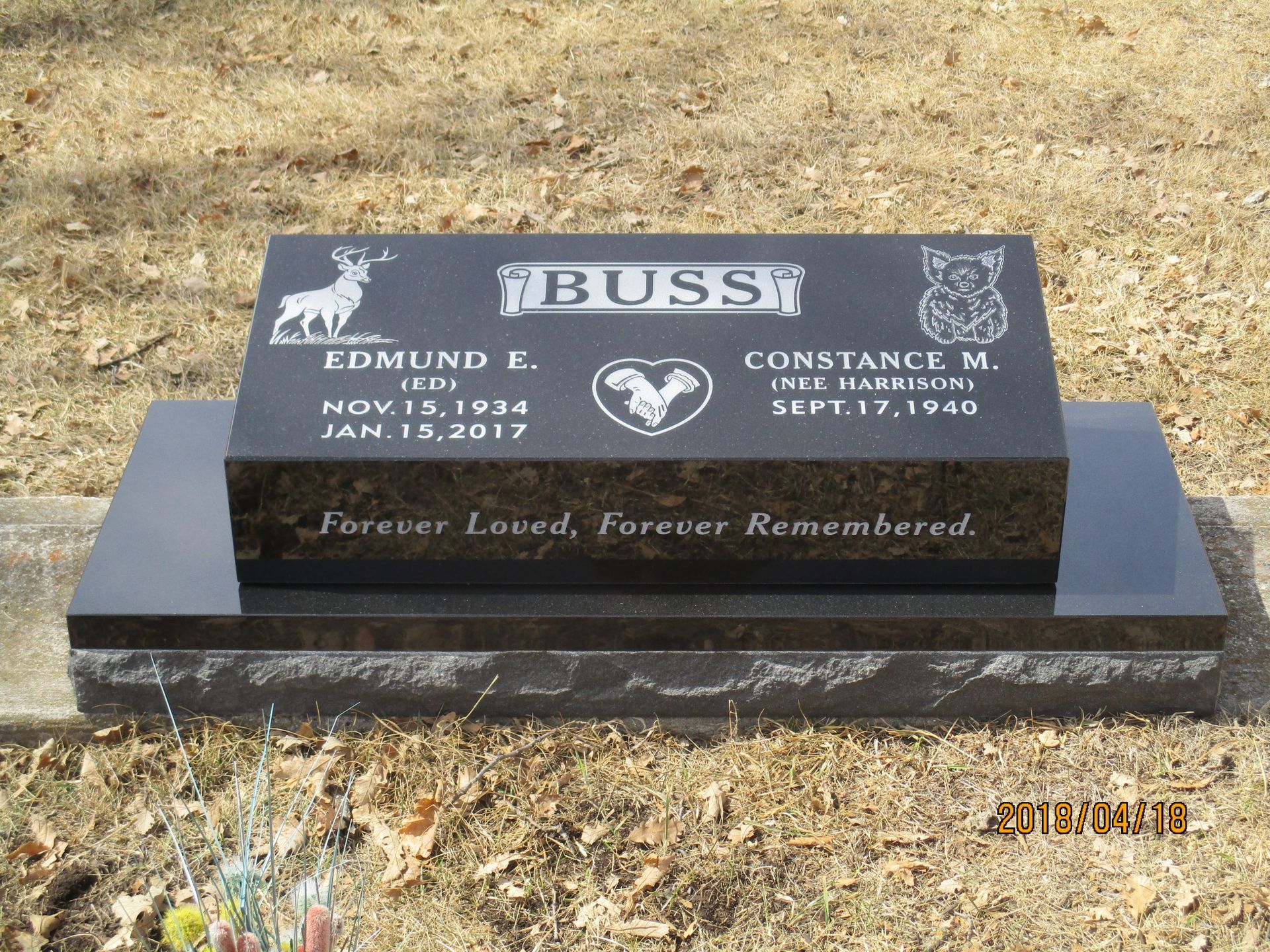 Black granite memorial headstone on dry grass with engraved names and “Forever Loved, Forever Remembered”