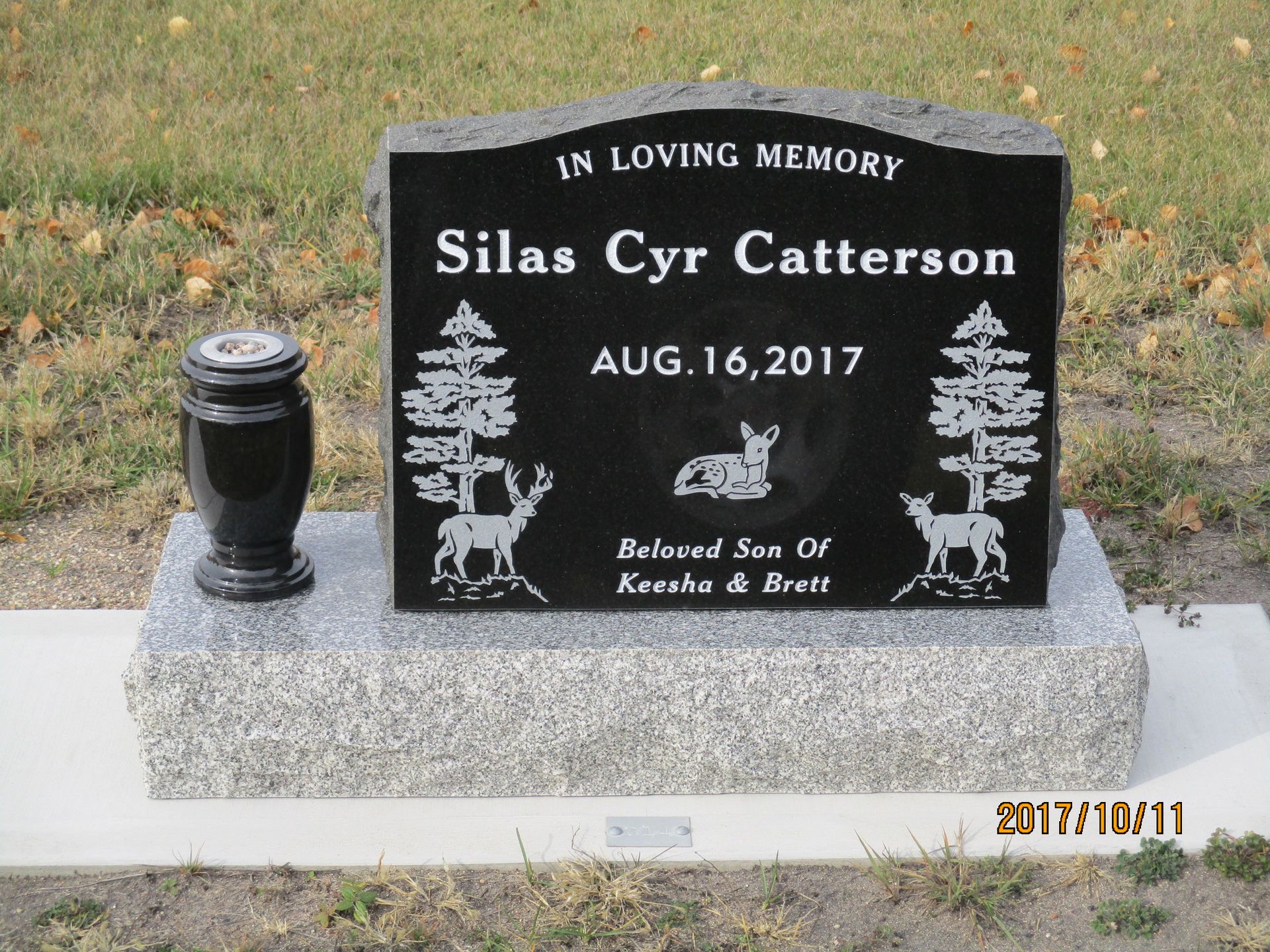 Black granite memorial stone on a concrete base in a grassy yard, with a small black urn beside it.