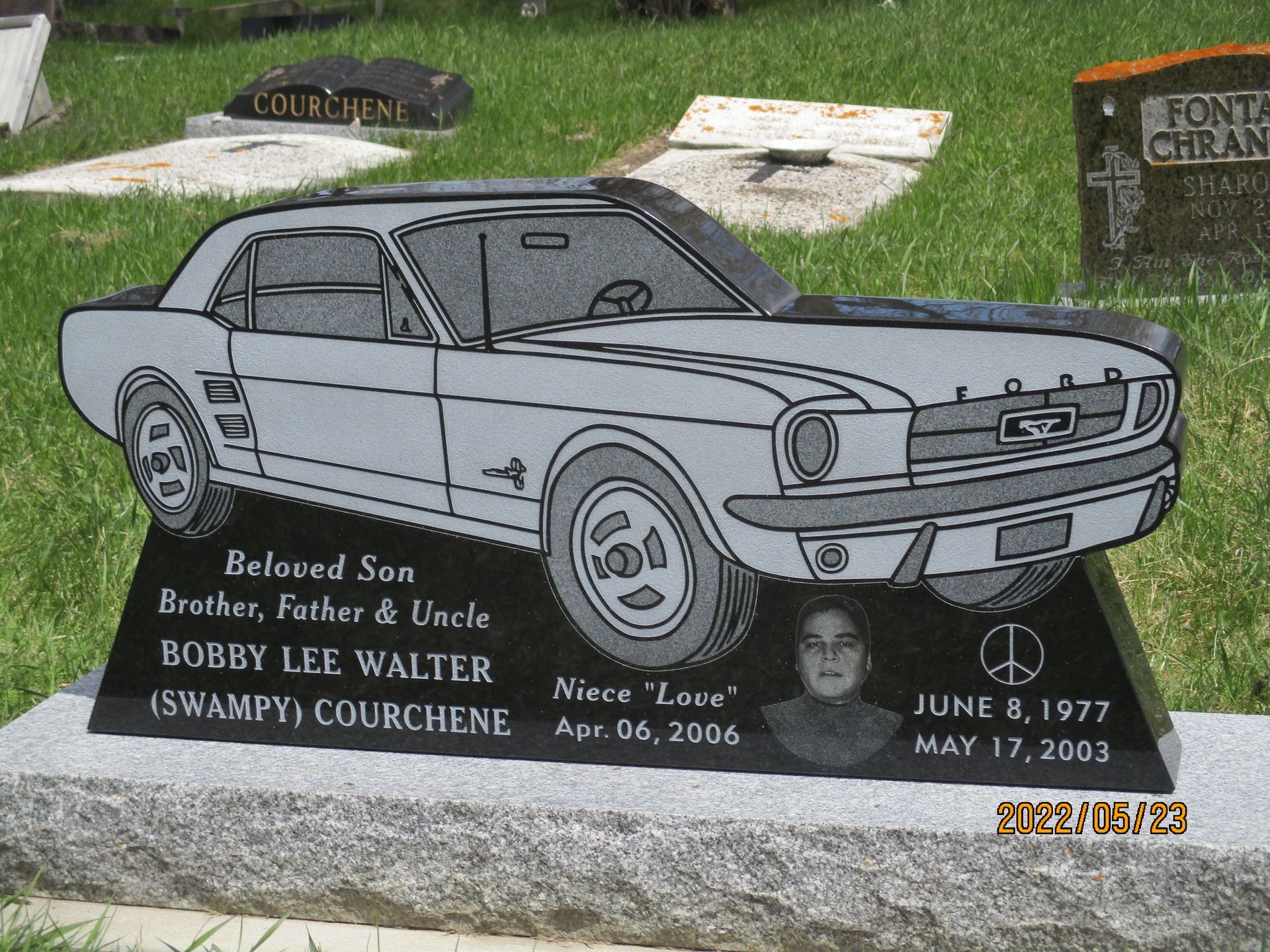 Gravestone engraved with a classic Ford Mustang, names, and dates in a cemetery.