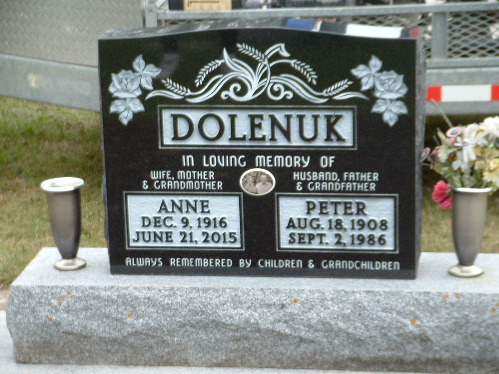 Black memorial headstone with flowers, engraved “DOLENUK,” in a cemetery.