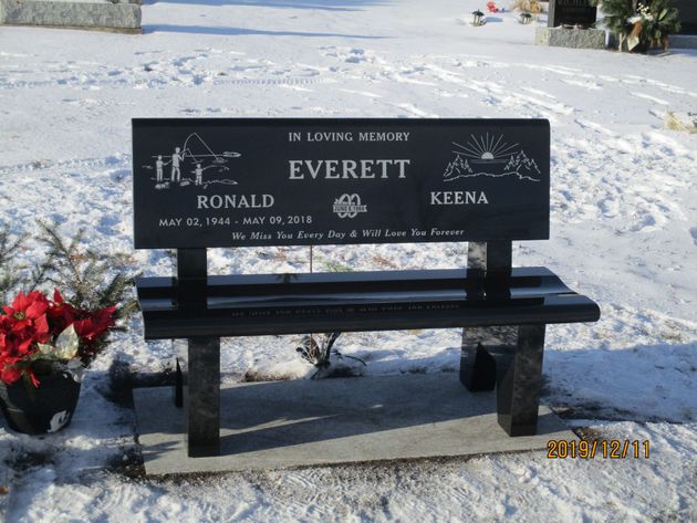 Snow-covered memorial bench with engraved plaque and flowers at a cemetery.