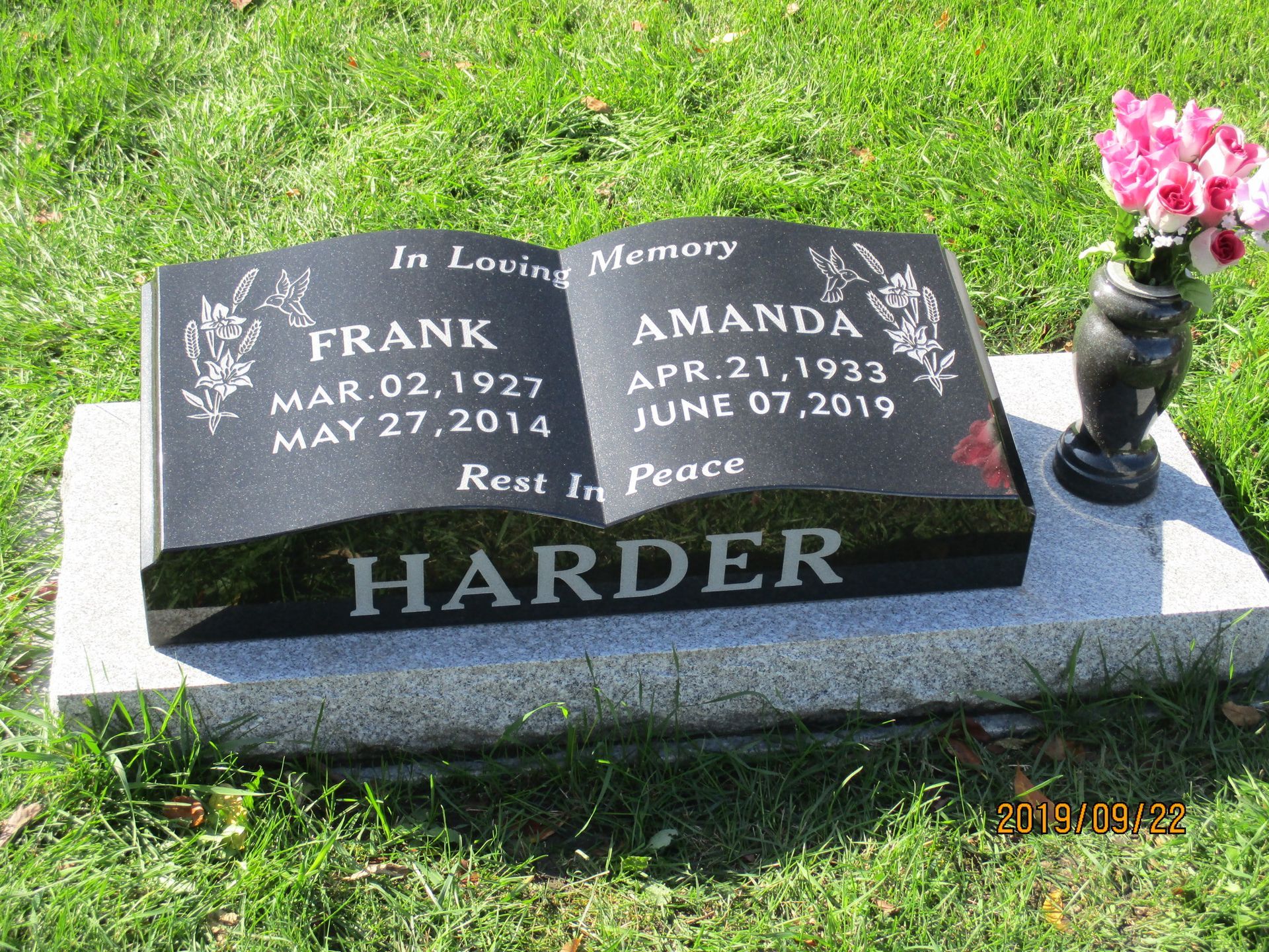 Black granite memorial headstone on a grave with pink flowers in a vase on the right.
