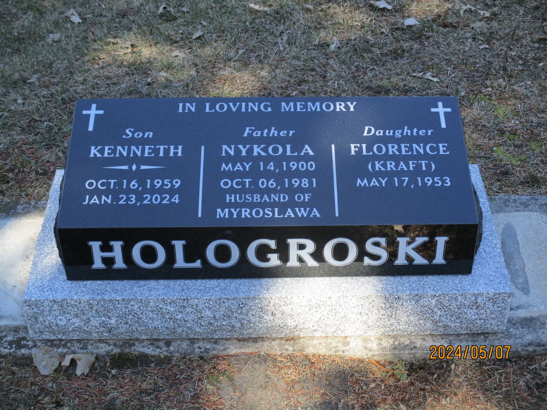 Memorial gravestone with blue plaques, crosses, and the name “HOLOGROSKI” on a cemetery marker