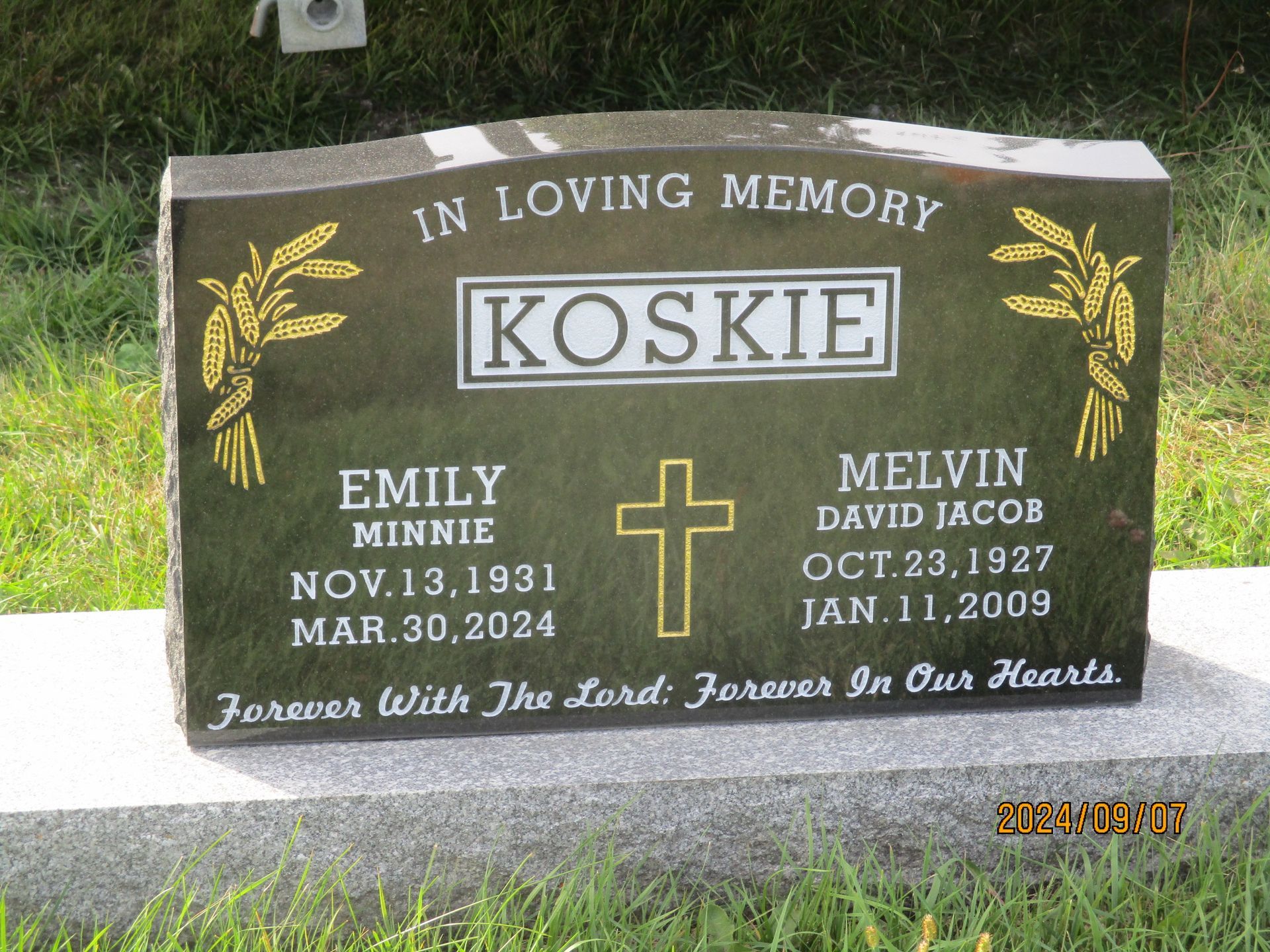 Black granite memorial headstone with gold lettering, cross, and angel designs in a grassy cemetery.