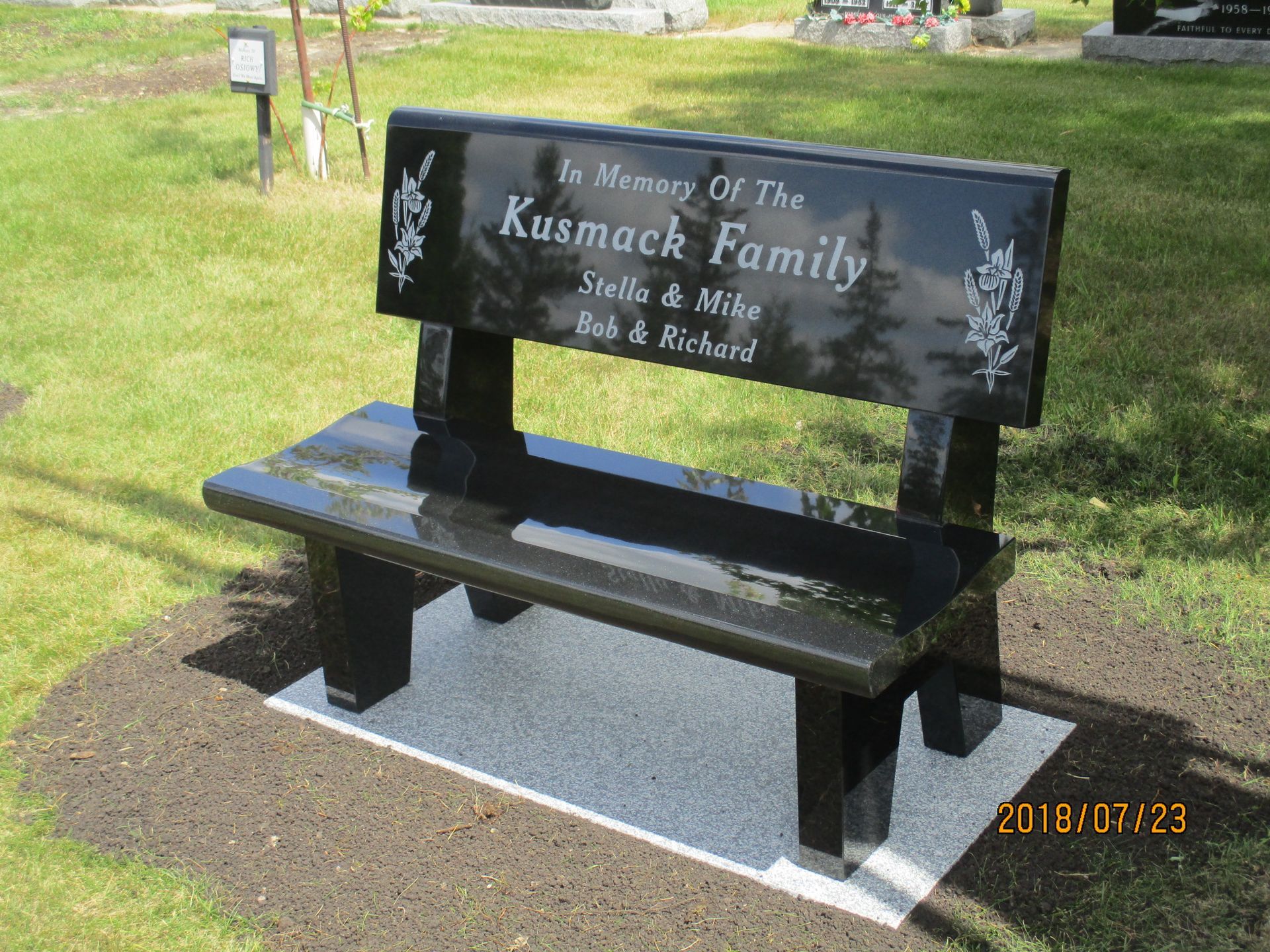 Black memorial bench for the Kusmak family with engraved text, set on a paved pad in a grassy area.