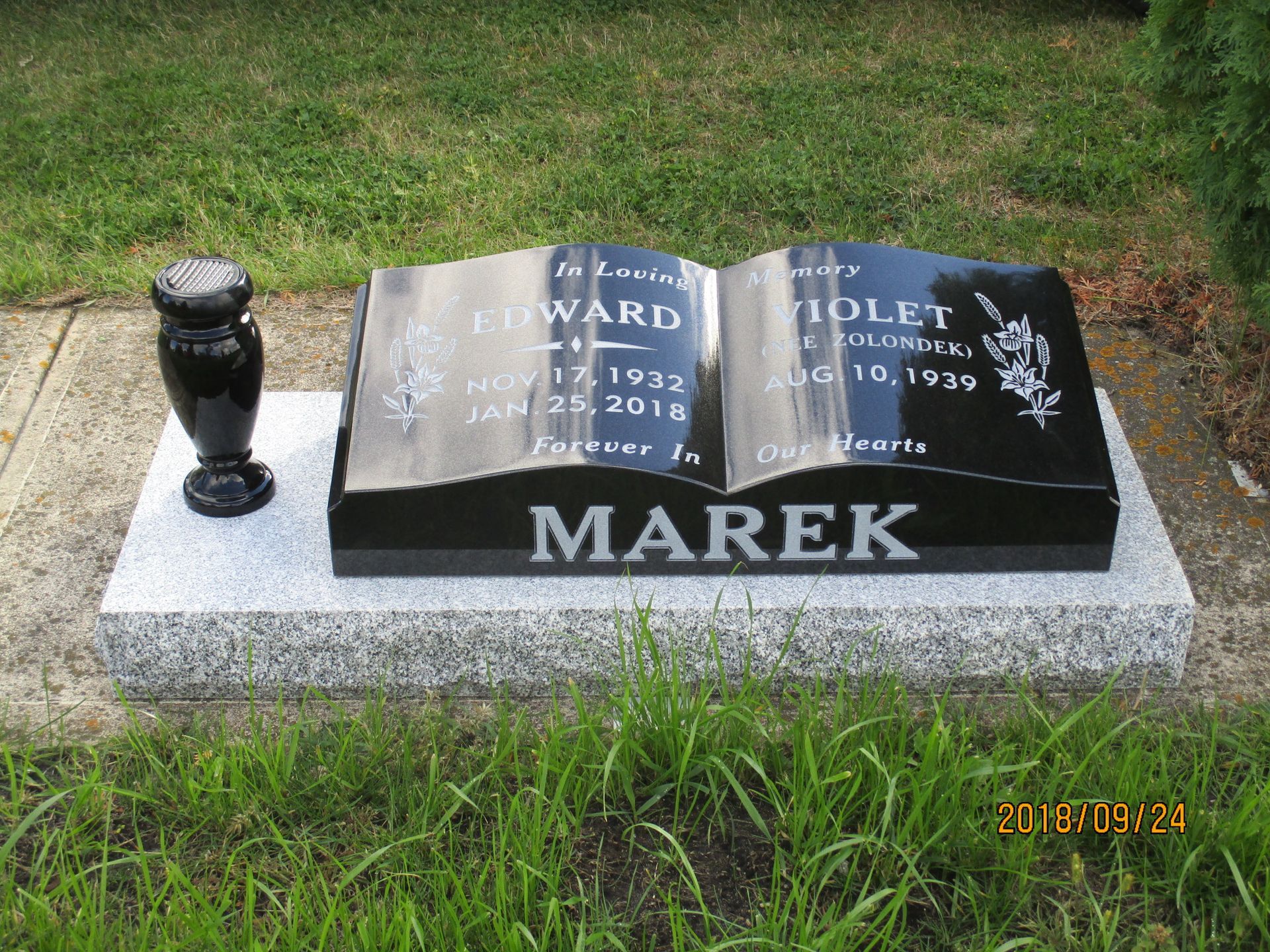 Black gravestone with polished plaque and urn on a concrete base in a grassy cemetery, marked MAREK