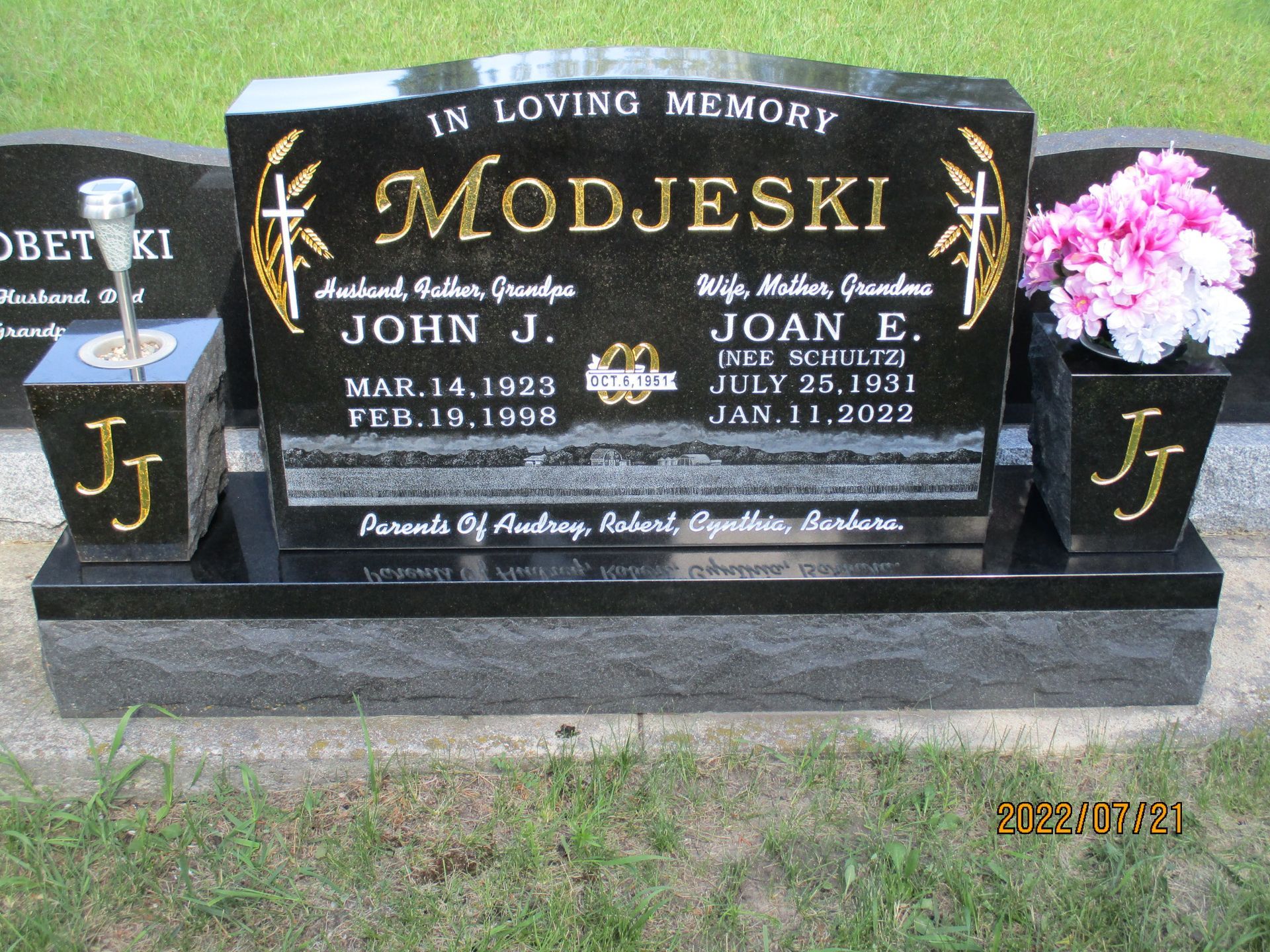 Black memorial gravestone with gold lettering and pink flowers, reading “Modjeski” and family names.