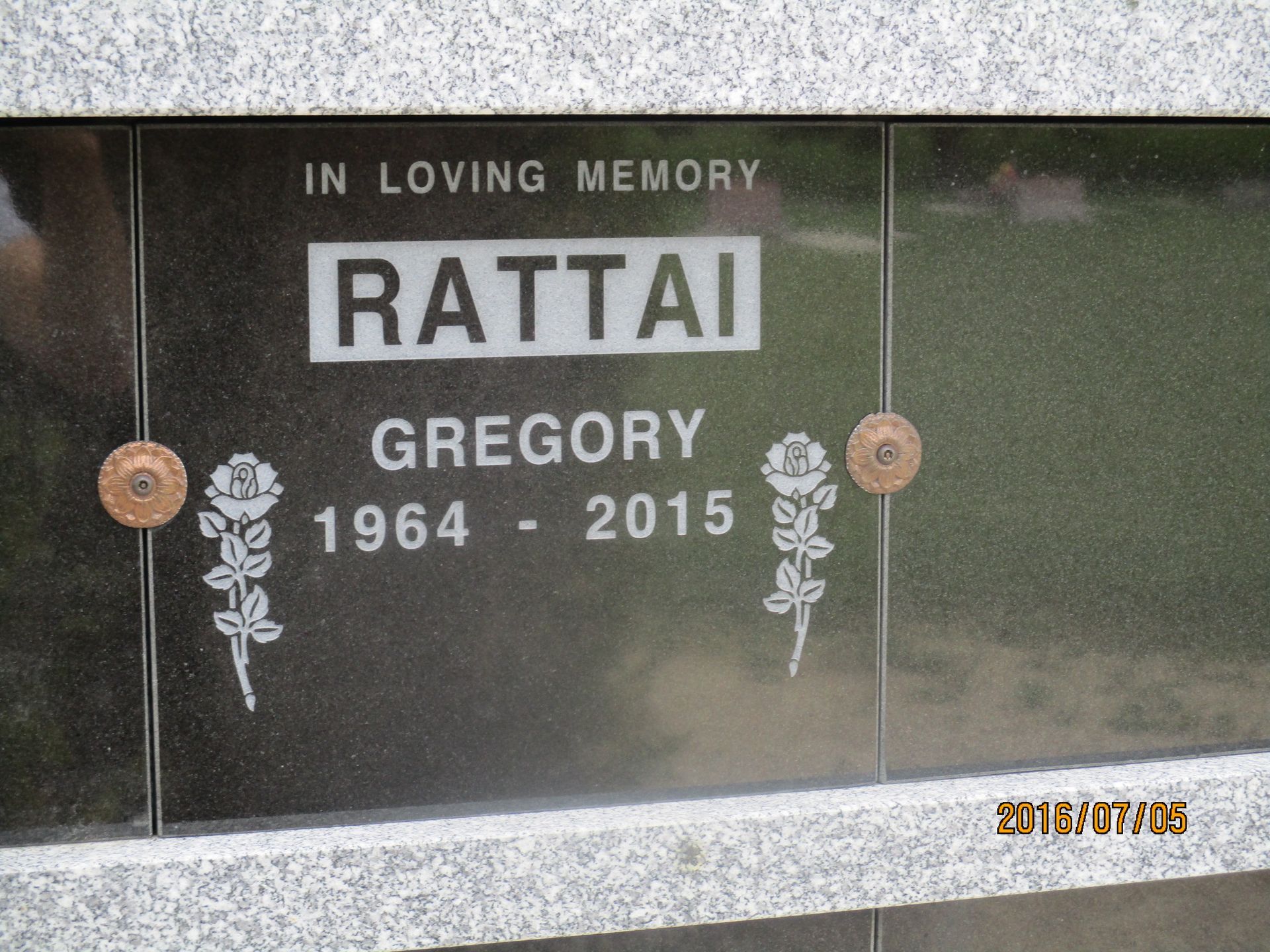 Black memorial plaque for Gregory Rattai, 1964–2015, with white lettering and floral accents.