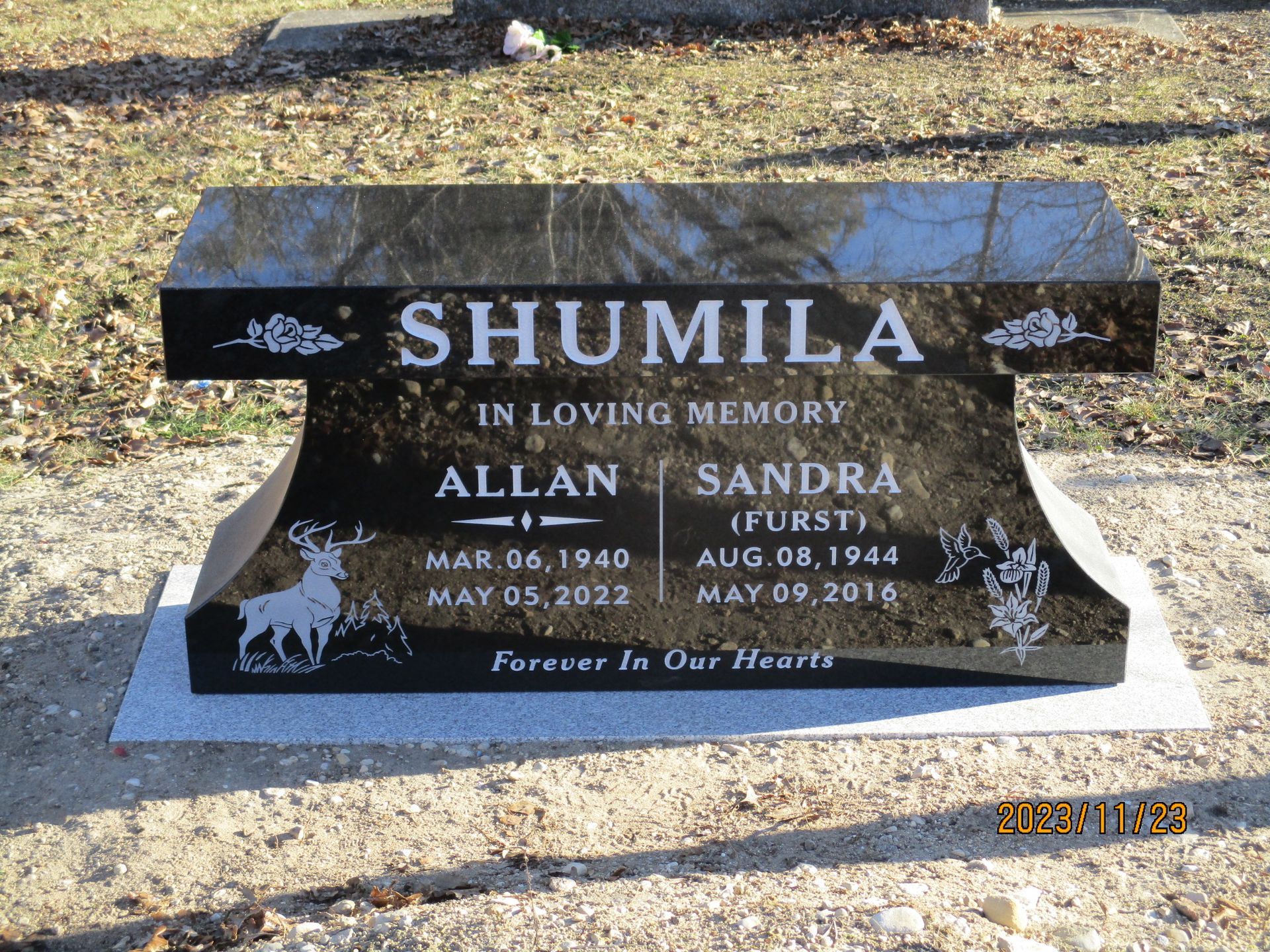 Black granite memorial headstone for Allan and Sandra Shumila in a grassy cemetery, with engraved names and dates