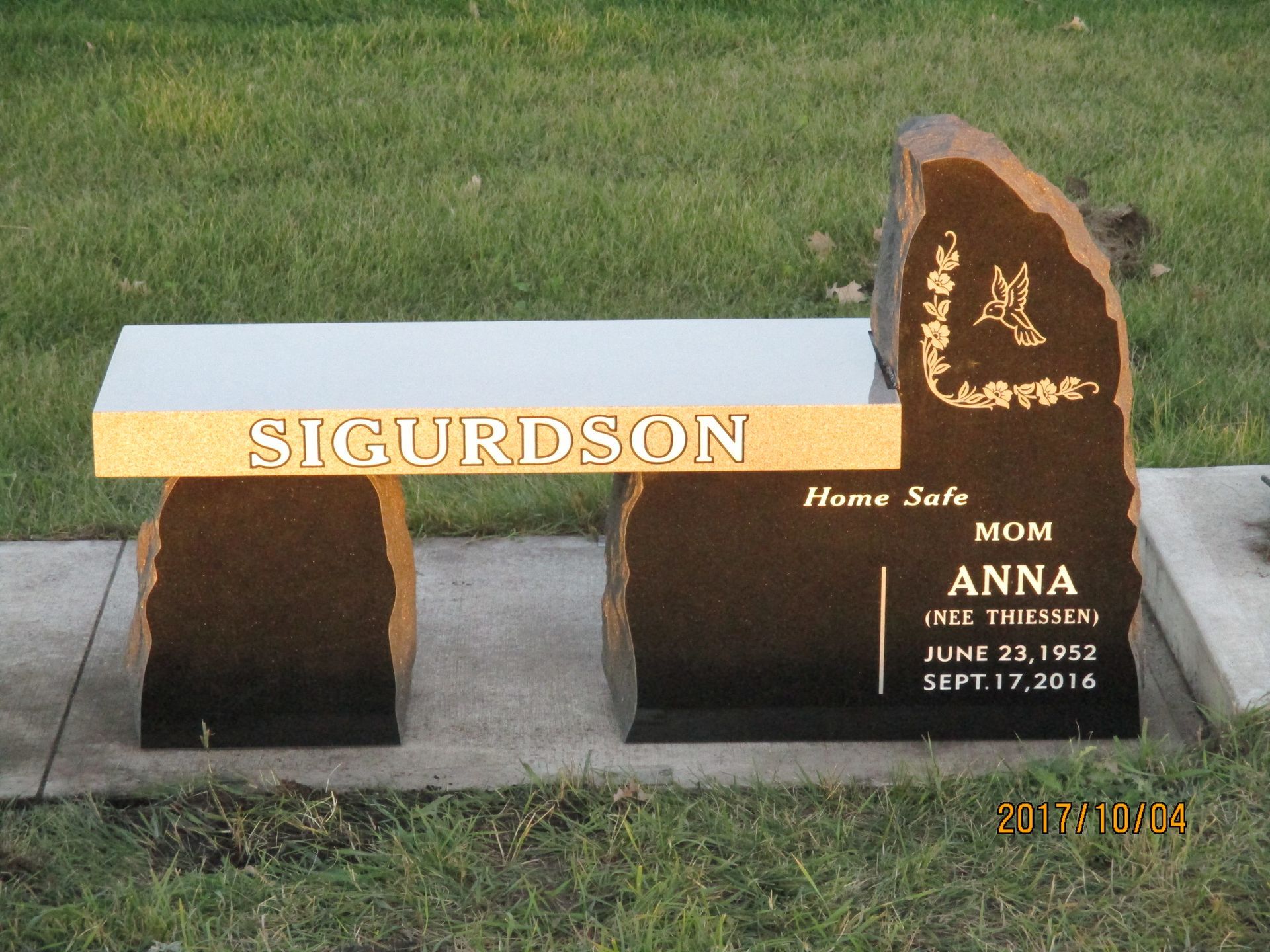 A memorial bench with the name SIGURDSON and a black-and-gold memorial plaque in a grassy cemetery setting