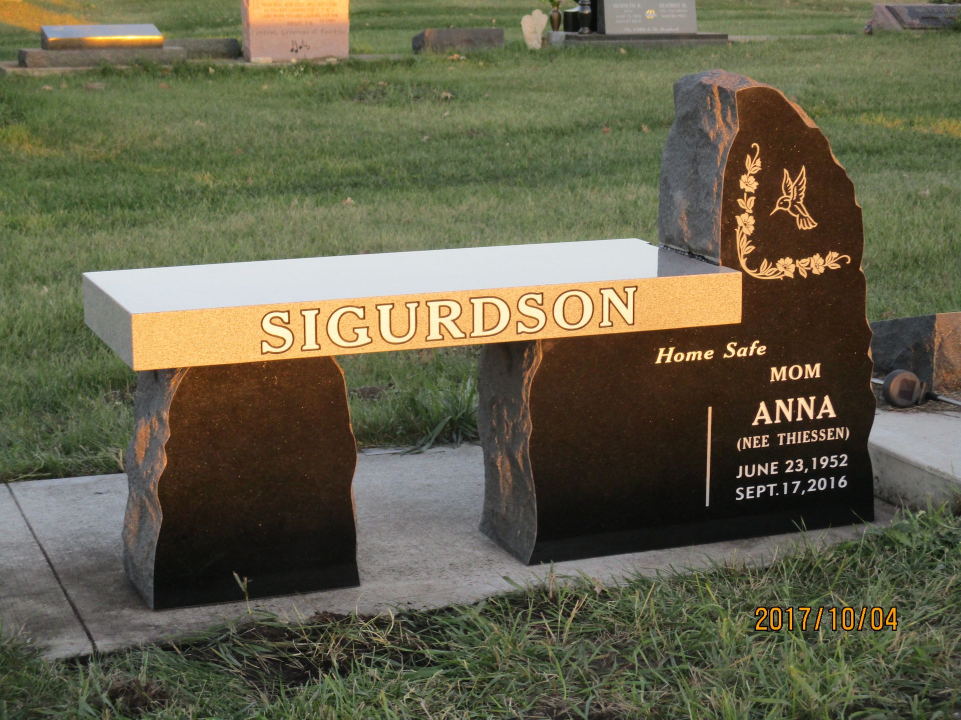 Memorial bench and plaque at a grassy cemetery, inscribed “SIGURDSON” and “Anna.”
