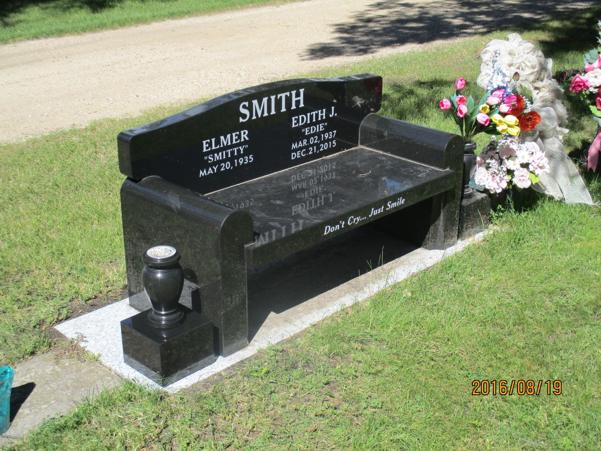 Black memorial bench gravestone with flowers beside it in a cemetery lawn