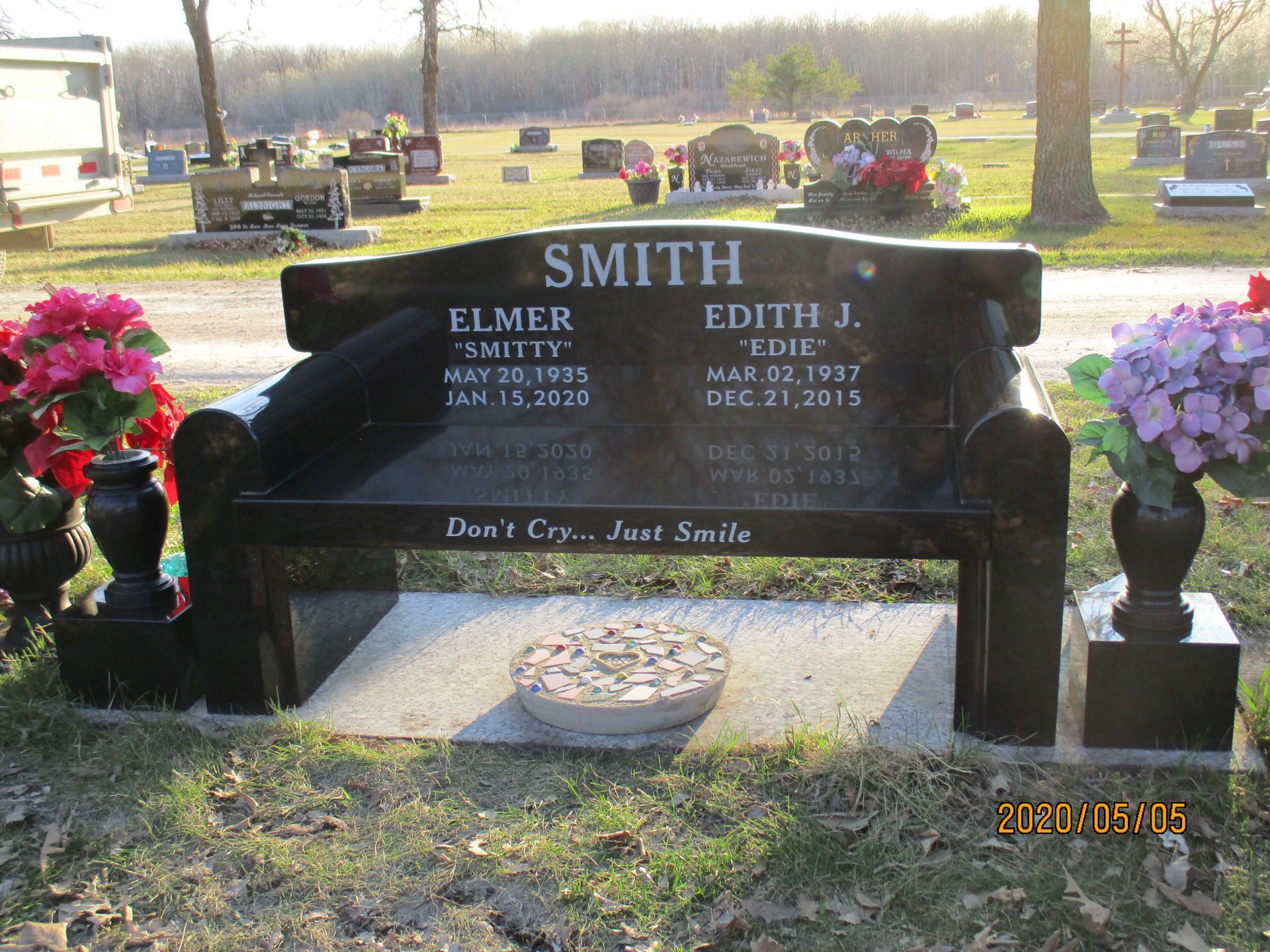 Gravestone with flowers on both sides in a sunlit cemetery; SMITH engraved on the black monument.