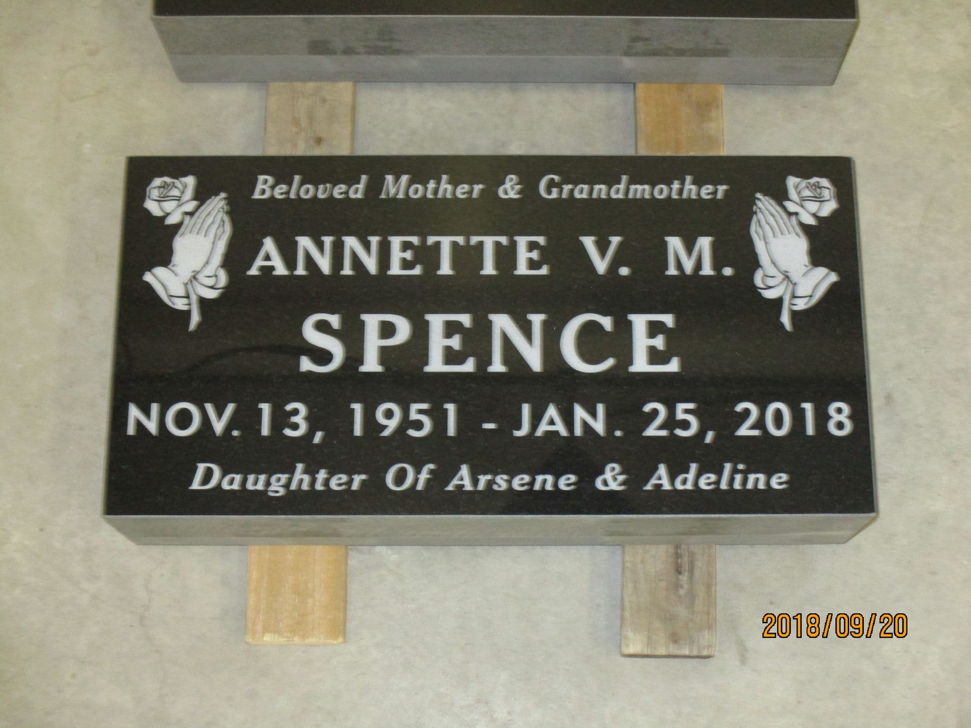 Black memorial plaque for Annette V. M. Spence with dates, on a wall with flowers engraved at the corners