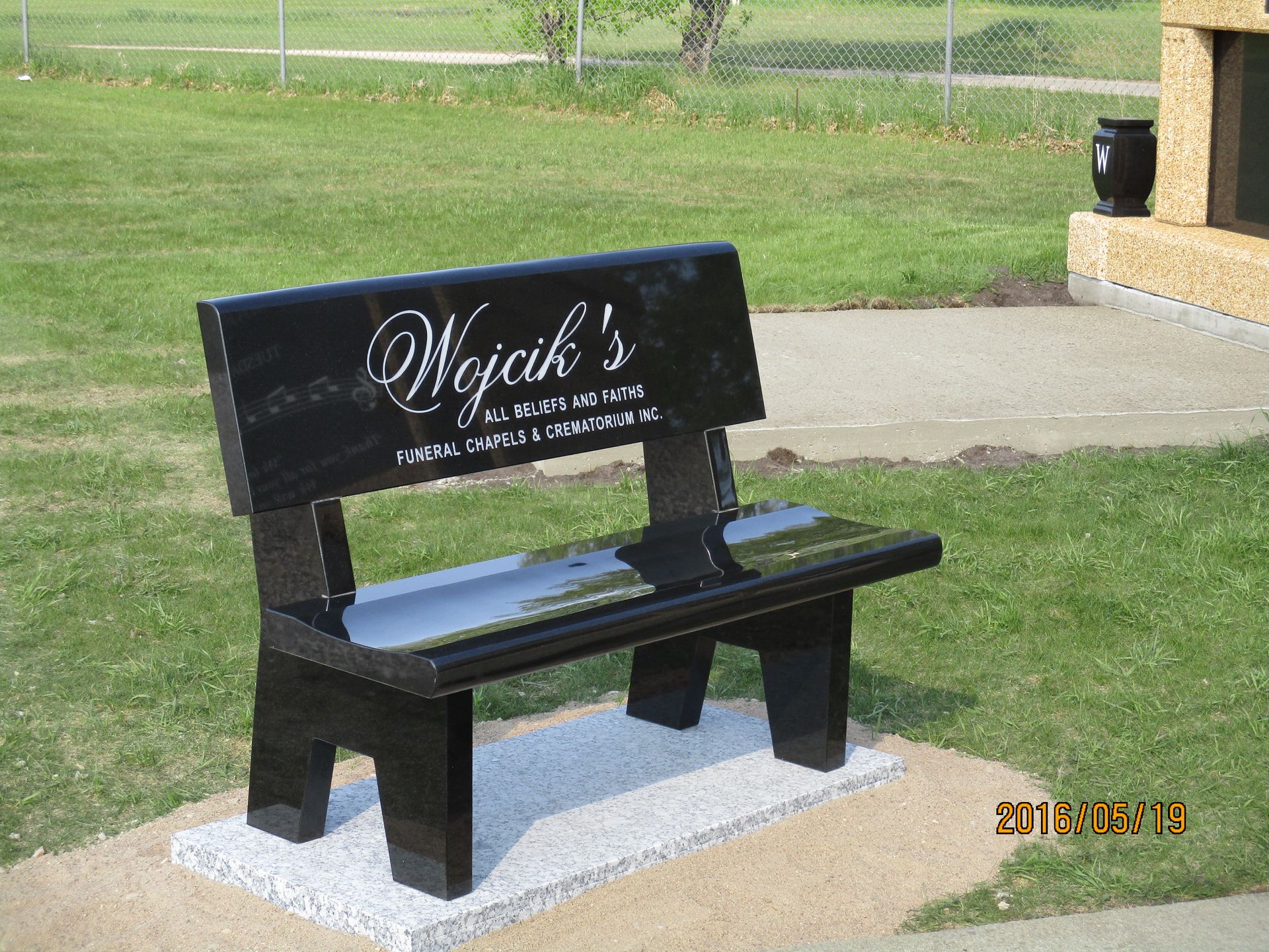 Black memorial bench on concrete pad in a grassy park near a sidewalk and building.