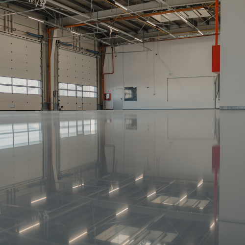 A large, empty industrial space with a shiny, reflective gray floor, white walls, and overhead lighting.