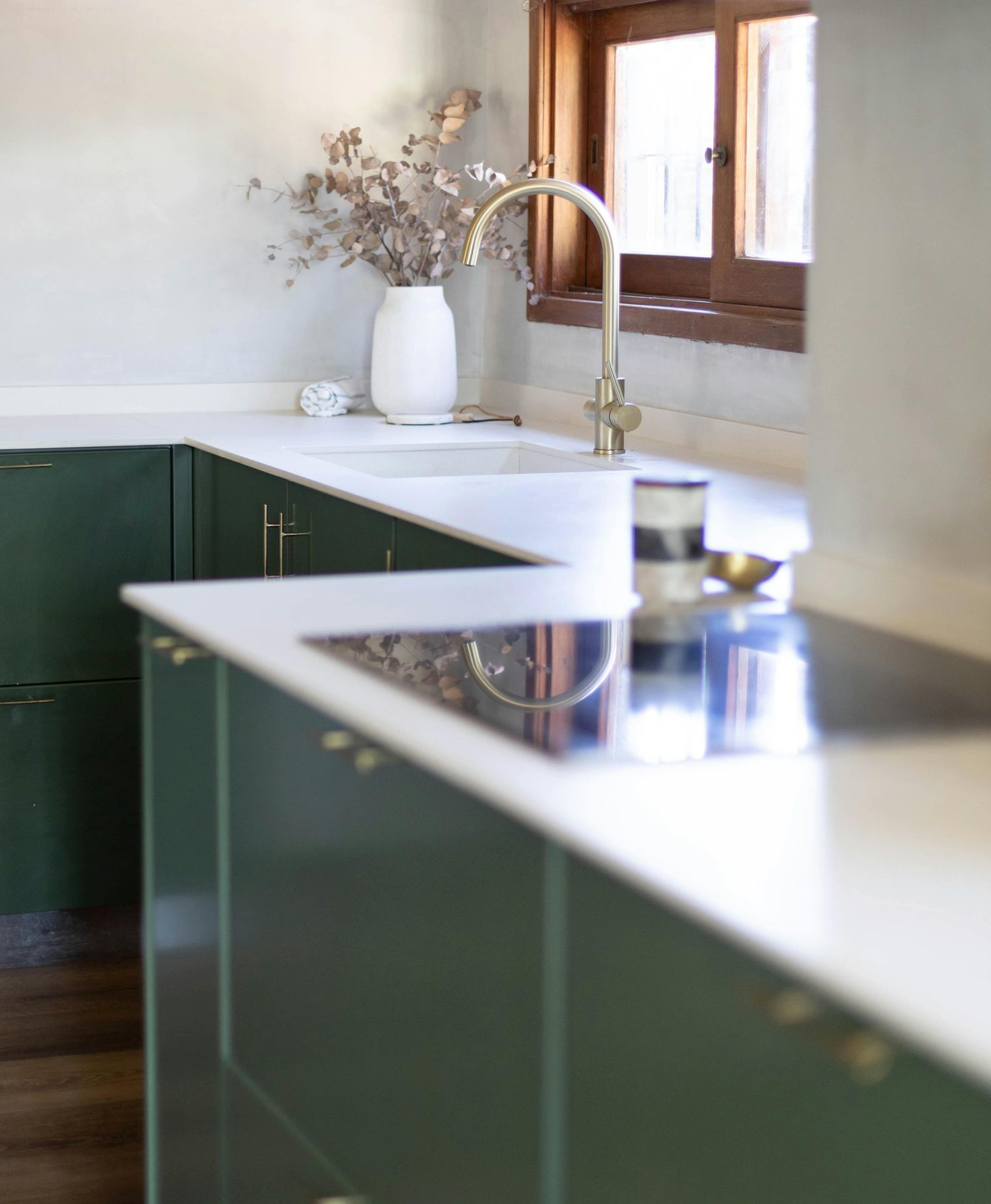 Green kitchen cabinets with white countertops, gold faucet and a vase of dried flowers.