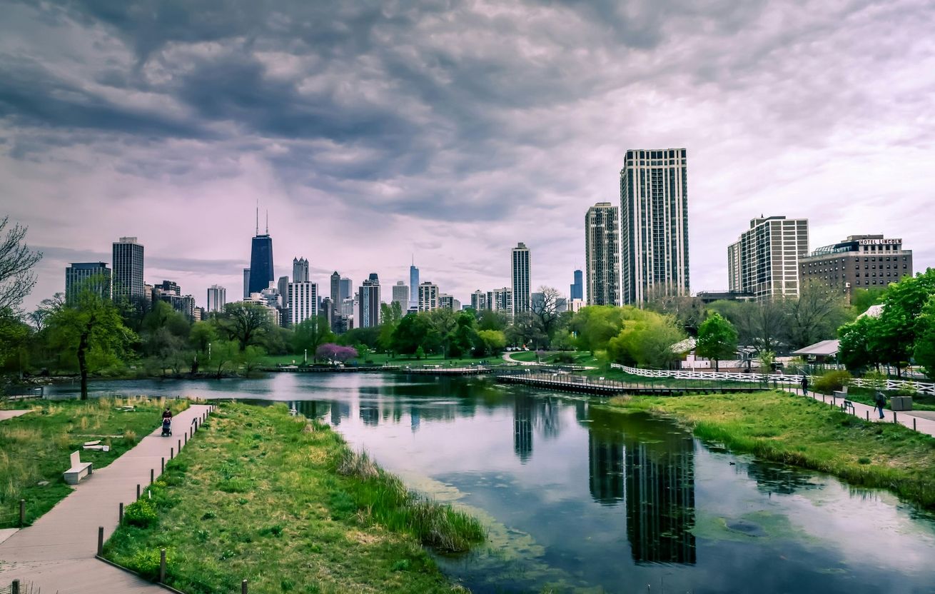 Chicago skyline view from a park, reflecting in calm water, under a cloudy sky.