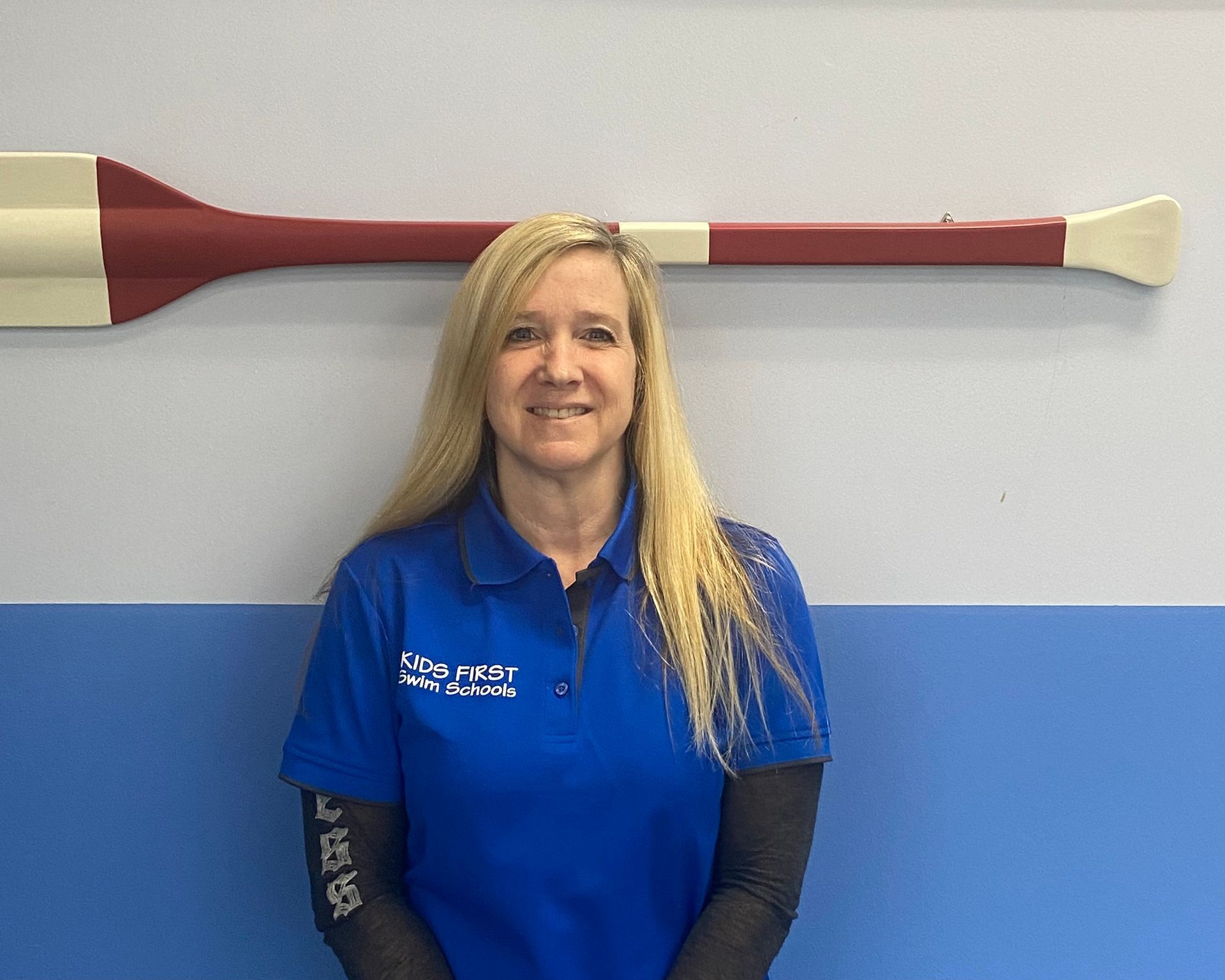 A woman in a blue shirt is standing in front of a large oar on a wall.