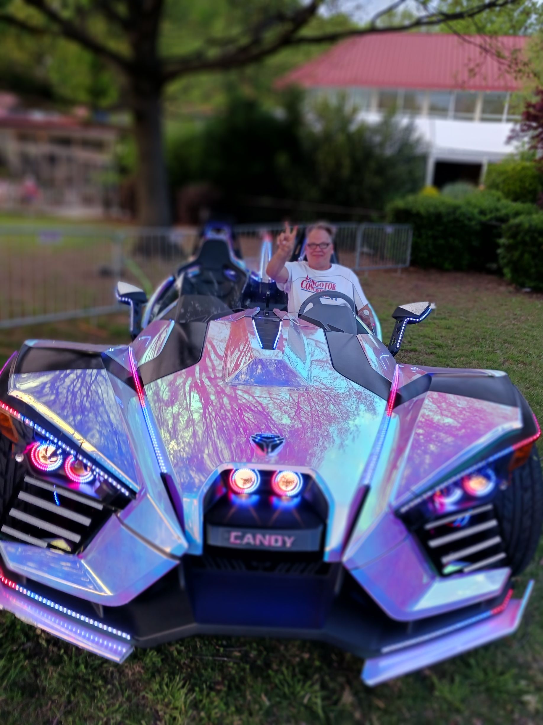 Iridescent three-wheeled Can-Am with two people seated, parked outdoors near a building.
