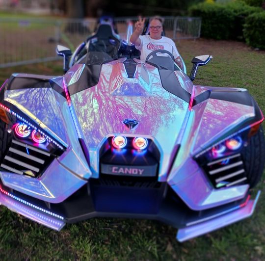 Person posing behind a colorful, neon-lit three-wheeled sports car outdoors