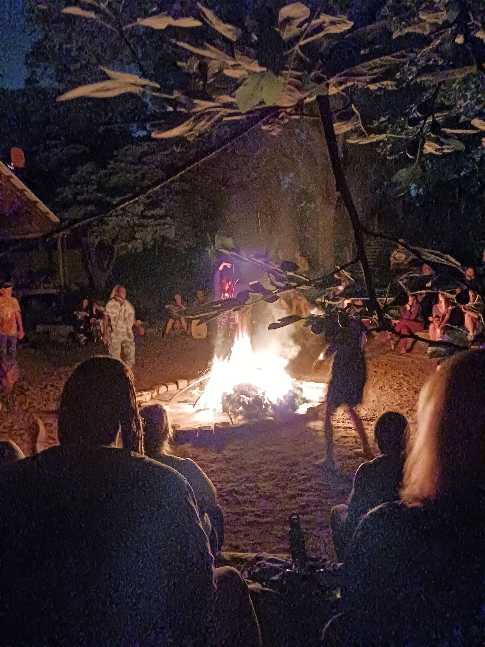 People gathered around a bright campfire at night in a wooded outdoor setting.