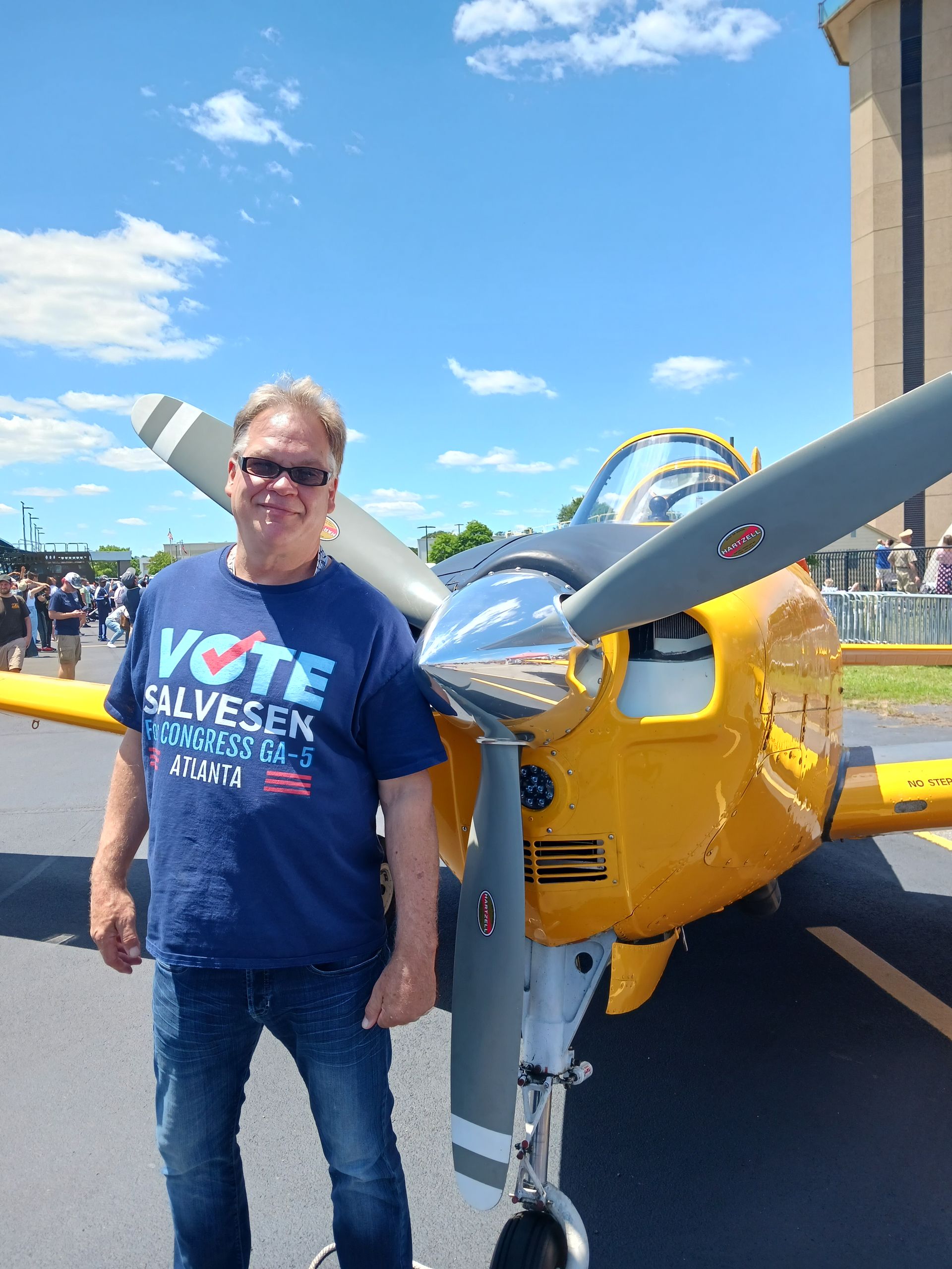 Man standing beside a yellow airplane with propeller, wearing a blue shirt and sunglasses under a sunny sky