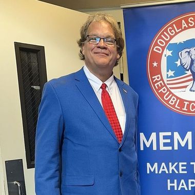 Man in a blue suit and red tie standing beside a Douglas County Republican banner.